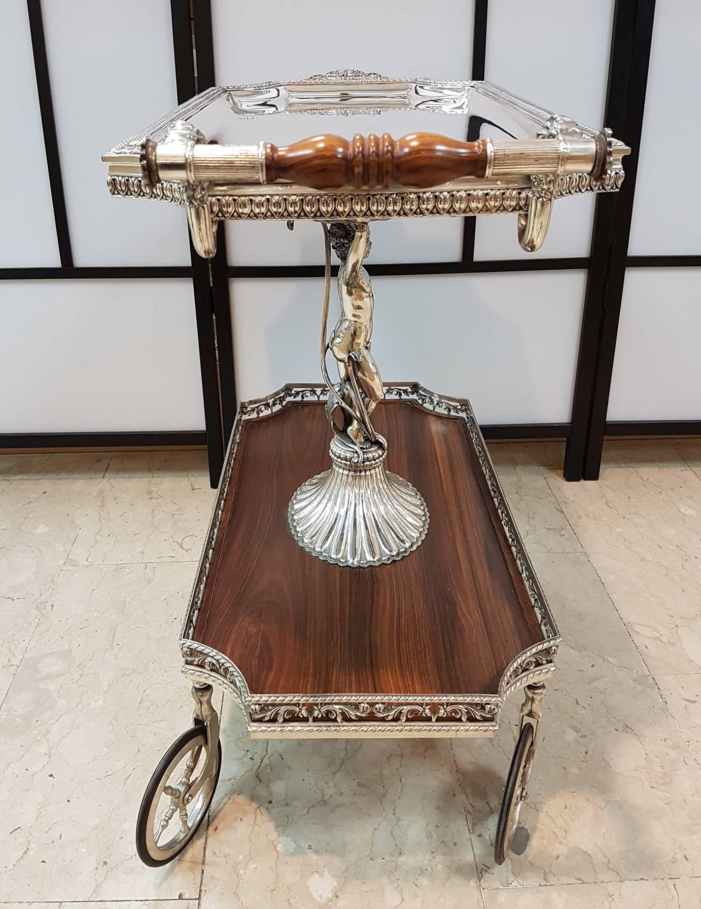 Solid silver trolley with tray on four wheels in silver.
The top is held up by a child in solid silver on fluted base.
The body is made of silver with two shelves the prized wood.
One handle with two biting lions to carry the trolley.
15,000 grams.