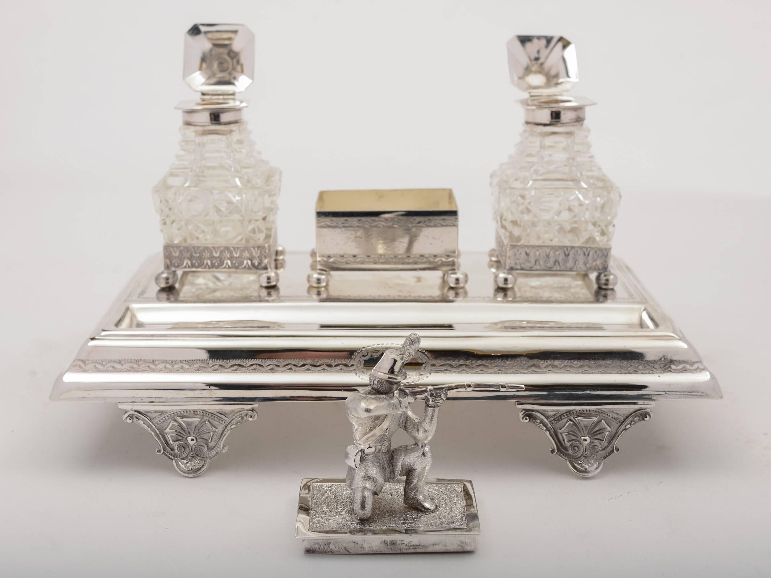 A stunning English Victorian silver plated ink stand with model of soldier in firing position crouched on stamp box in centre, flanked by X2 cut glass inkwells which have pagoda shaped tops. The stand sits on four embossed cast feet and there is