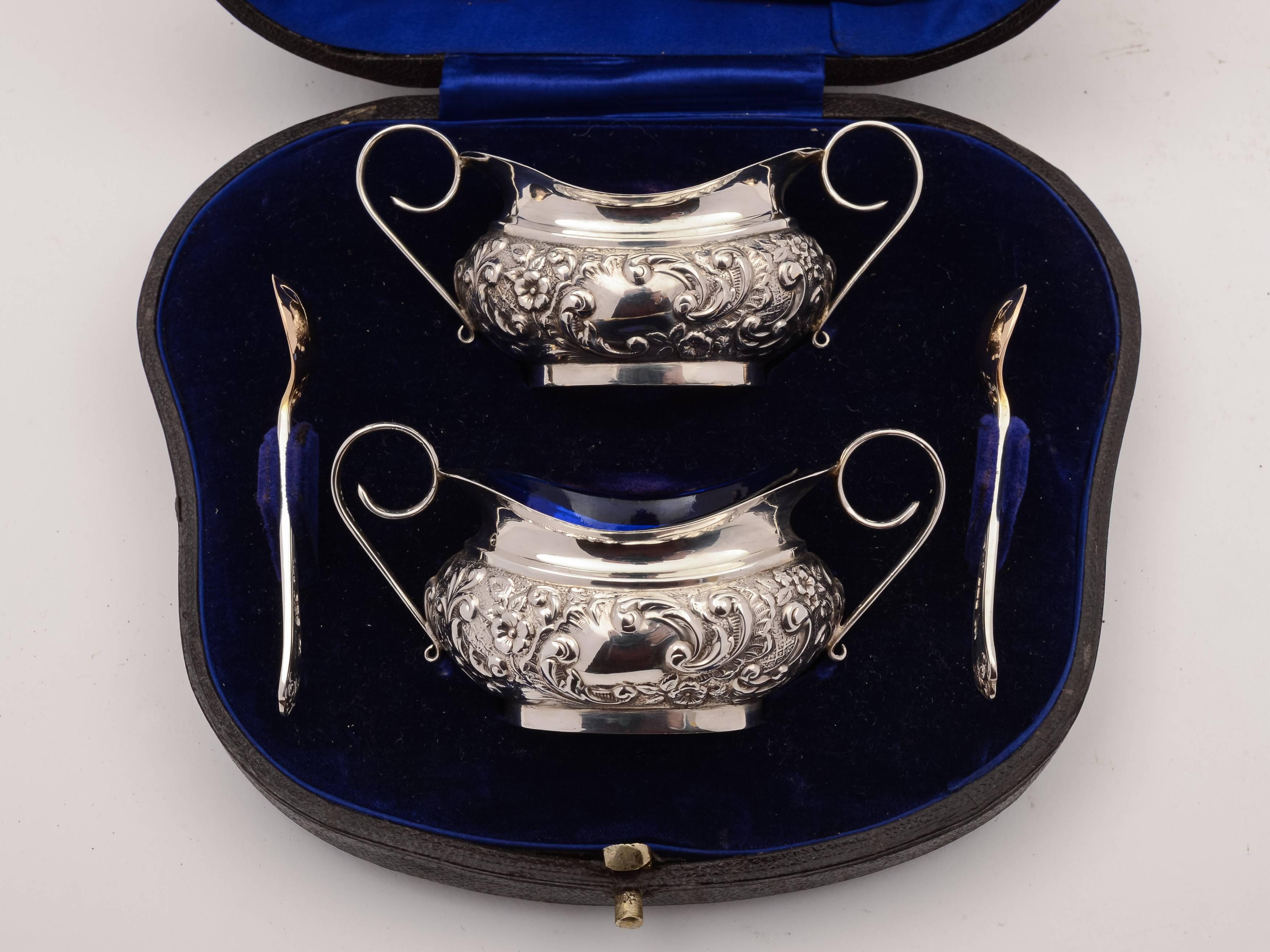 A lovely pair of English Edwardian silver salts with embossed decoration and scroll handles, come with silver salt spoons and original gilt interior with cobalt blue glass liners. Presented in original blue velvet and silk lined case. Hallmarked