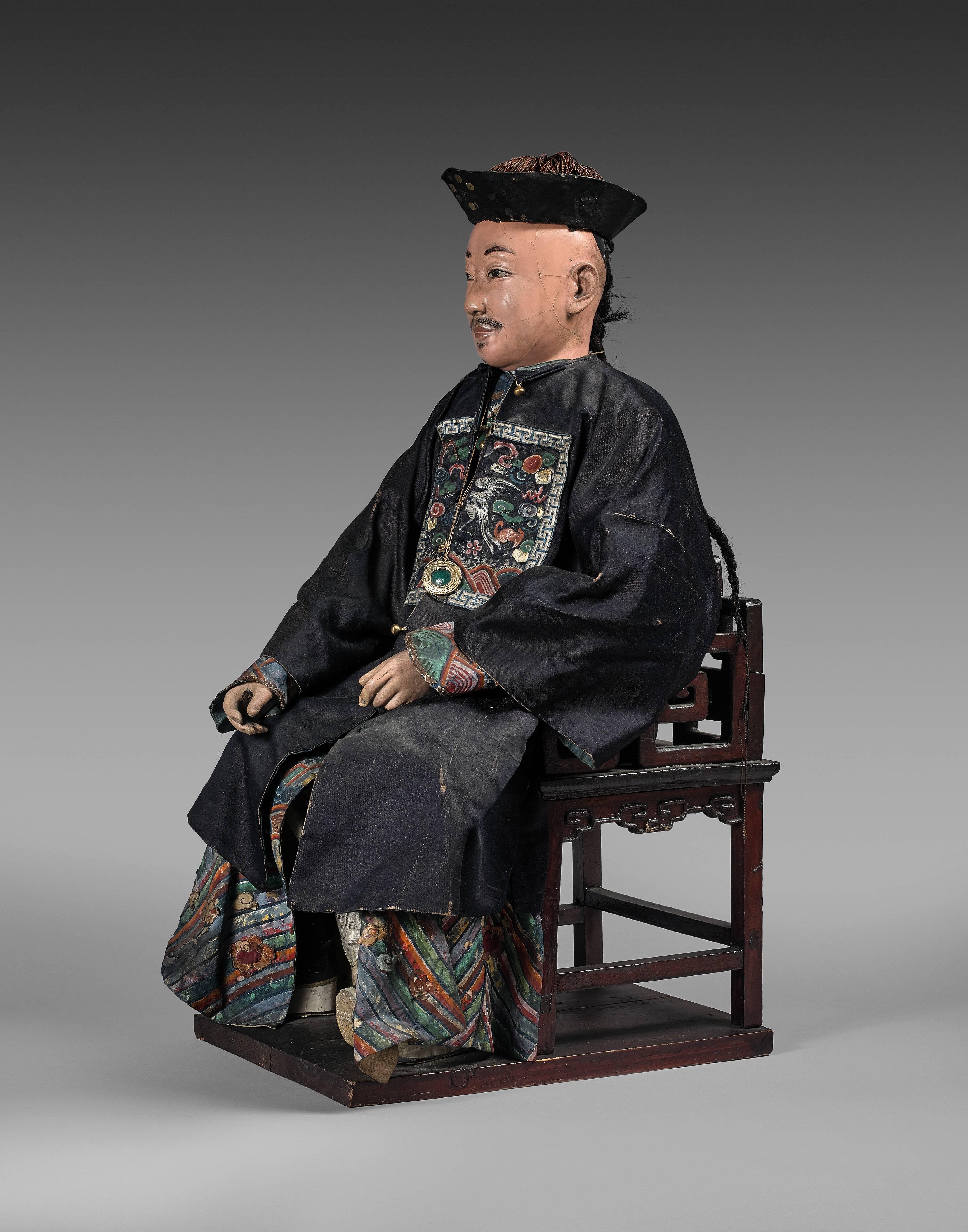 The Hong merchant seated on a Chinese style armchair, the different parts of the body articulated. The head and hands made of painted clay and carefully detailed. His eyes and eyebrows delicately painted, his hair in a long braid. The body made of