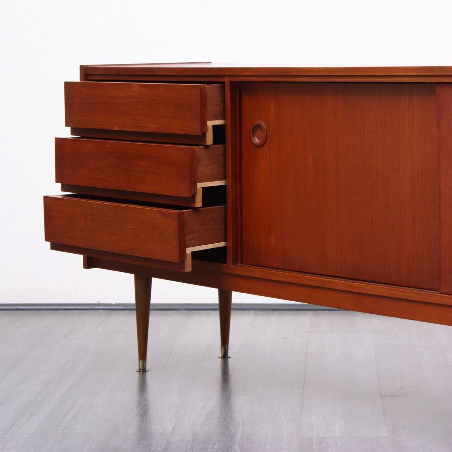Sideboard from the 1960s, Scandinavian design. Teak carcass with three drawers and two sliding doors with nice handles. Light bevelles feet with brass endings. Teak veneered, light chatoyant surfaces.Good condition with small traces of usage.