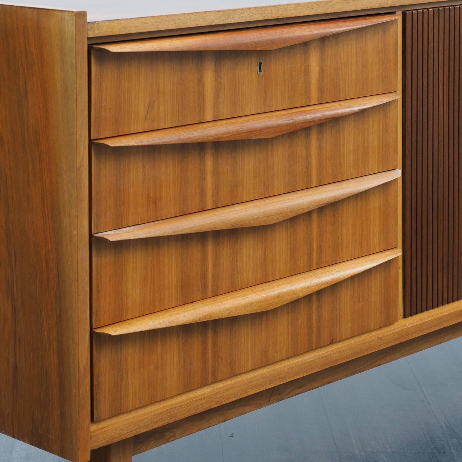 Rare 1950s sideboard with decorative structured doors. The appearance is compact and the board comes in the highly sought after length: 189cm. The sideboard is made detailed throughout with beautiful, molded solid wood-handles on the four drawers.