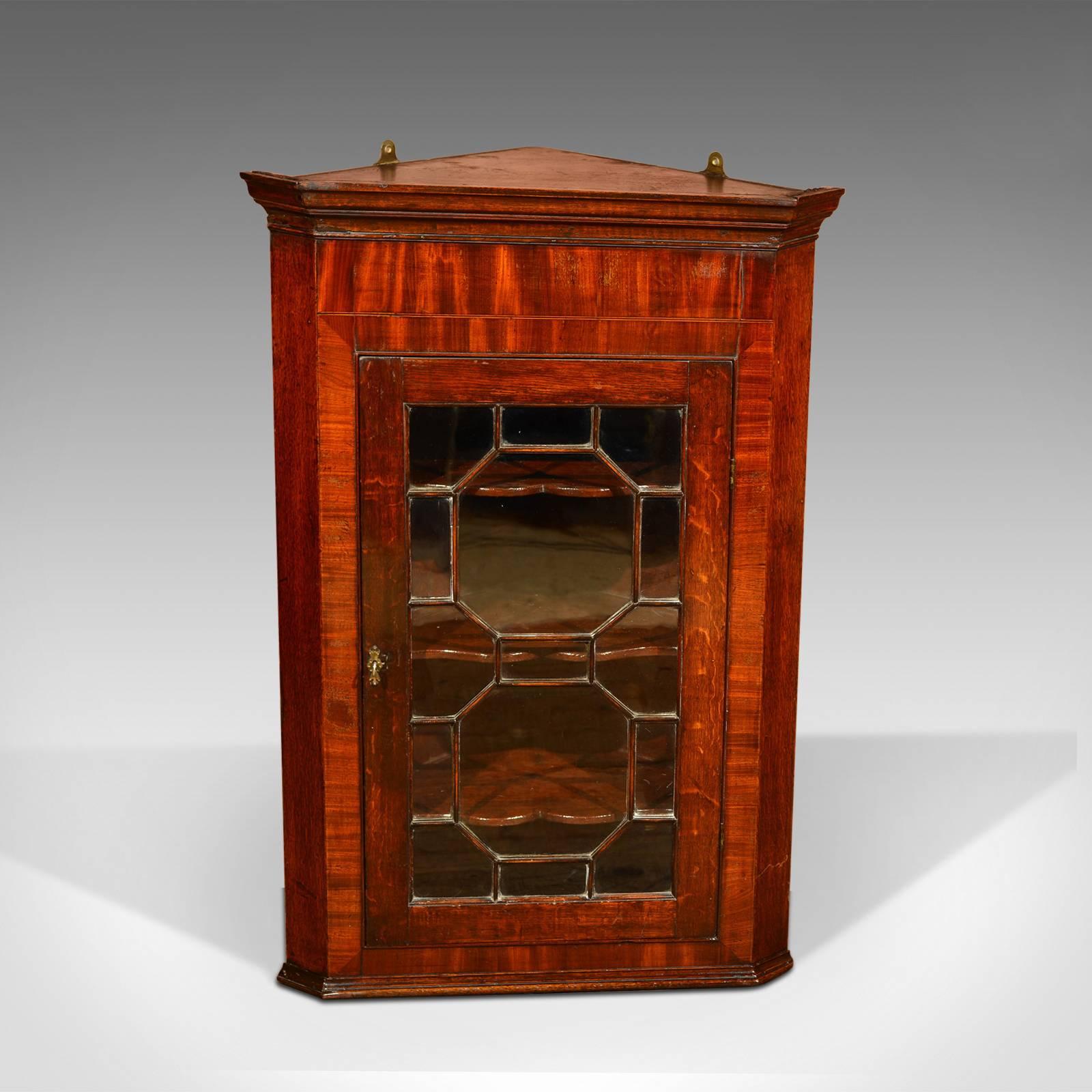 This is an antique, Georgian, hanging antique corner cabinet dating to circa 1800.

This mid-sized cabinet is constructed in oak with canted corners and offers added interest with the mahogany veneer framing around the door panel. The fifteen pane