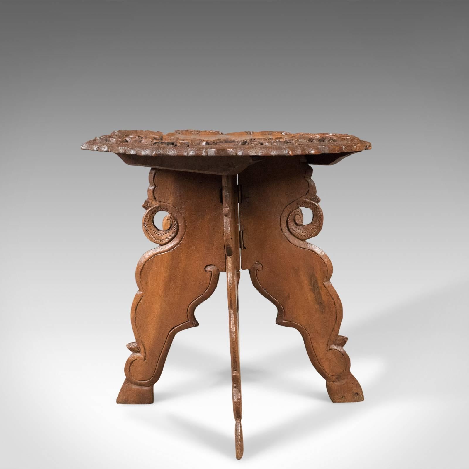 This is an attractive, carved teak, Campaign side table from the mid-20th century.

Central, ribbon edged tabletop with good grain detail
Broad, carved border displaying floral sprays
Outer shaped edge of octagonal form

Fixed legs splayed for