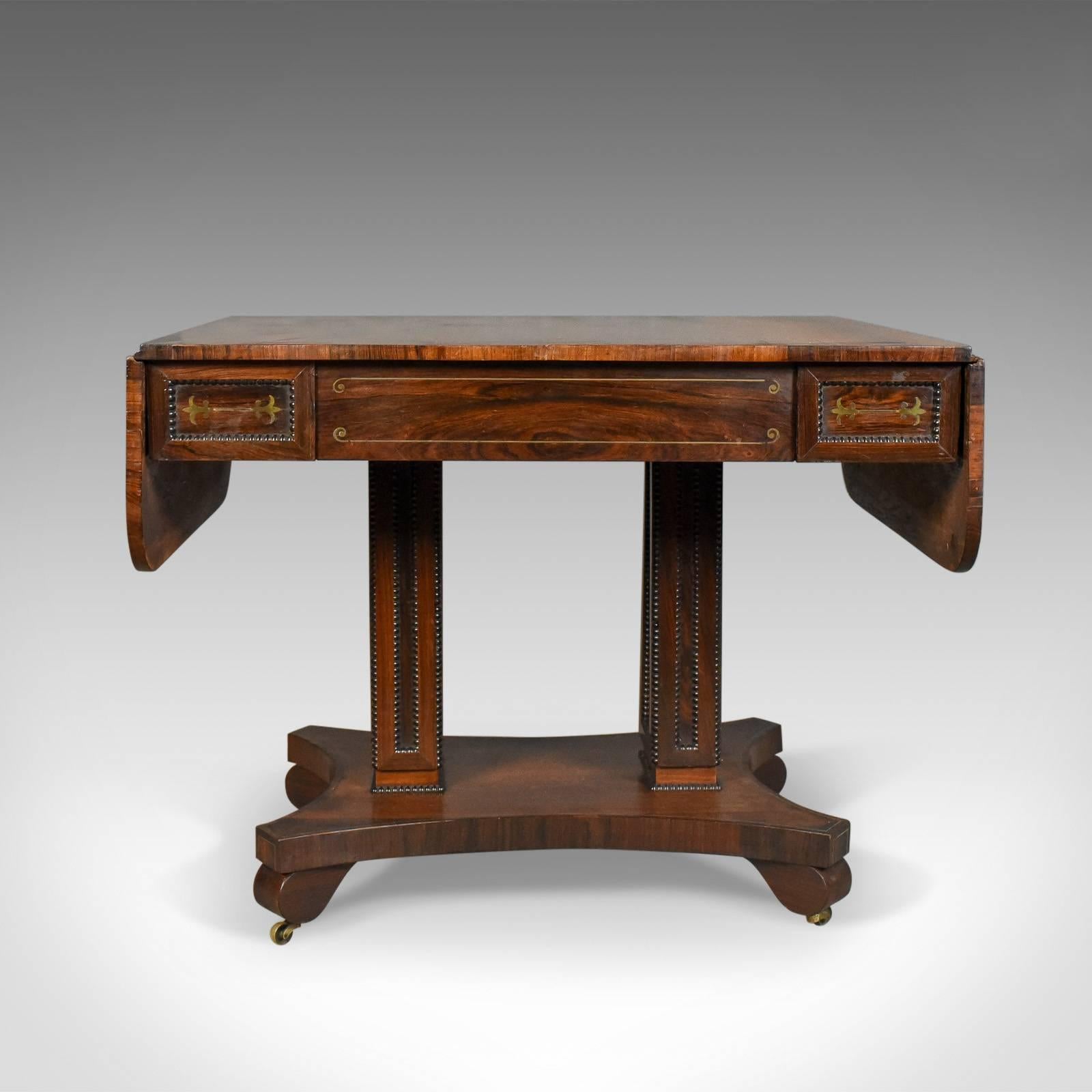 This is an antique sofa table in rosewood, an English Regency Pembroke table dating to circa 1820.

Superb color and grain interest in the well figured rosewood
Desirable aged patina and of quality craftsmanship
Highlighted and decorated with