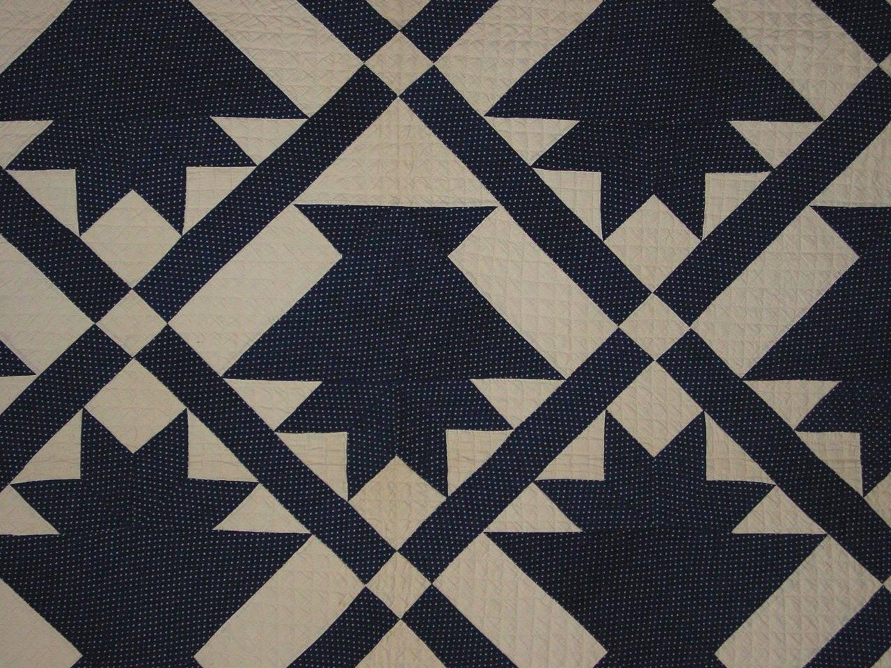 A graphic Folk Art patchwork quilt composed of blue cotton calico baskets on a white quilted ground. A cloth label at bottom left corner reads 