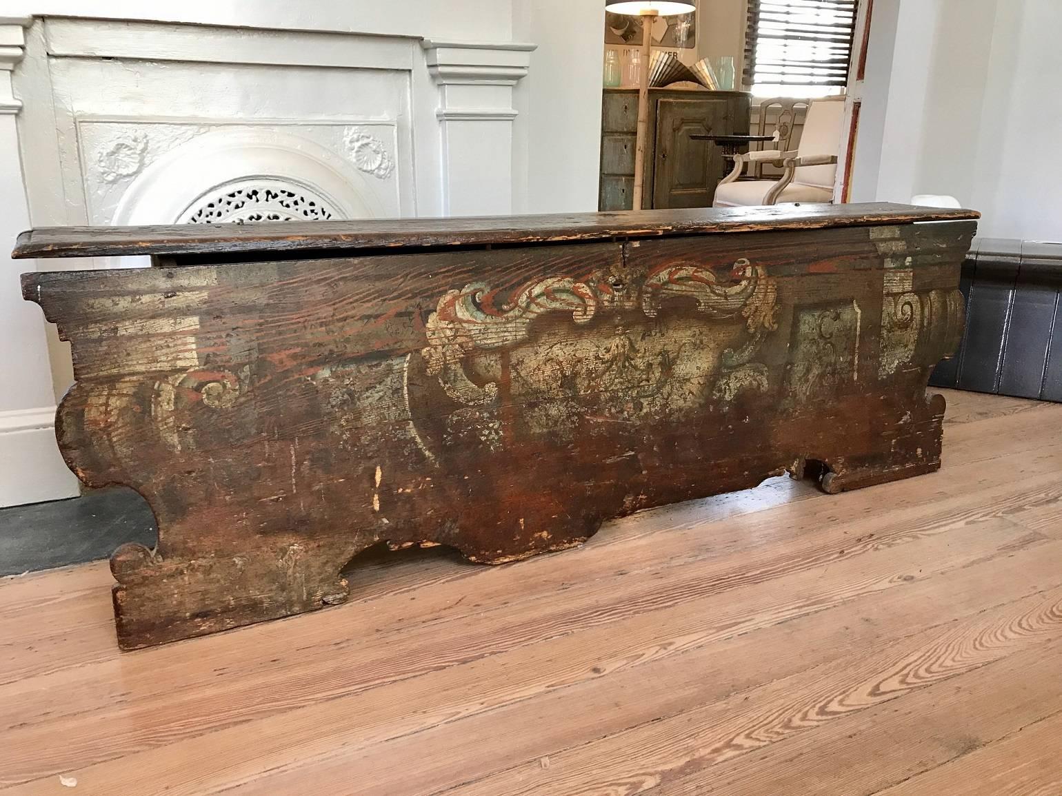 Wooden Italian bench with two storage sections beneath a hinged lid the front panel extending beyond the body of the cabinet at both ends; decorated with painted motifs in the classical taste suggesting column capitals flanking a central cartouche;
