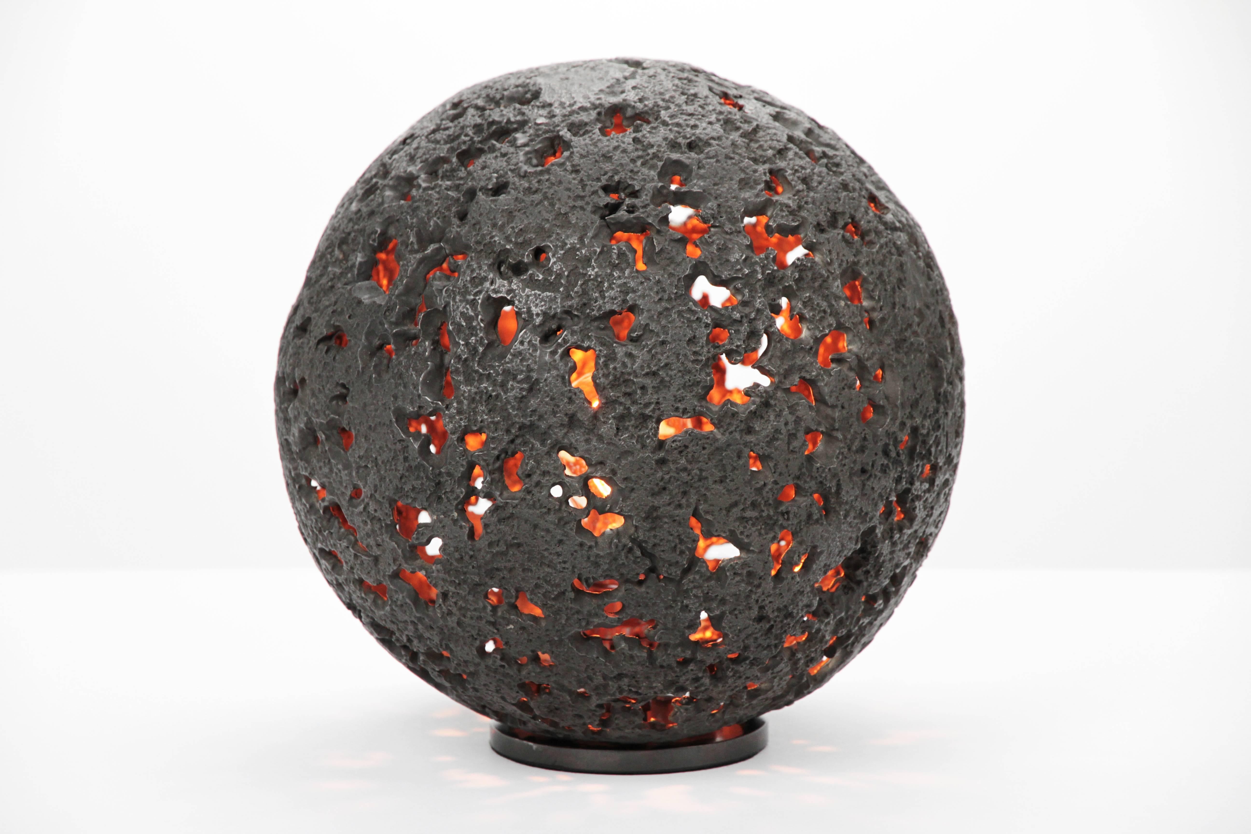 The Hot Planet Table Lamp is a sphere full of craters with a light source at the center symbolizing the core of energy within a planet.  The body is cast in bronze with a blackened patina. The patina brings out the texture in the sculpture.  The raw