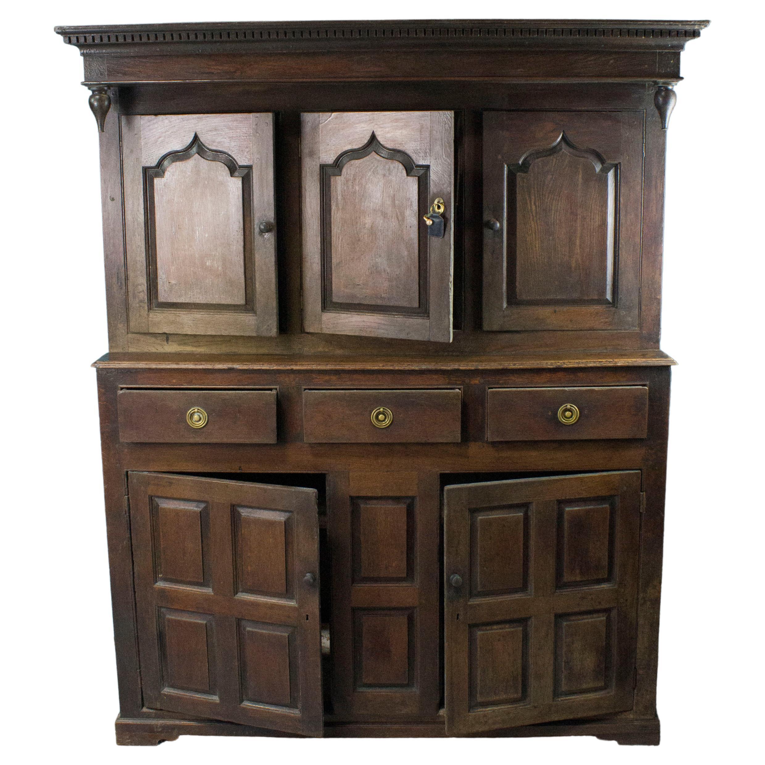 18th century oak sideboard opening with two doors with panels in the lower part with wooden buttons and three belt drawers with brass catches. The upper part of the cupboard, set back slightly, has three molded doors and a projecting cornice