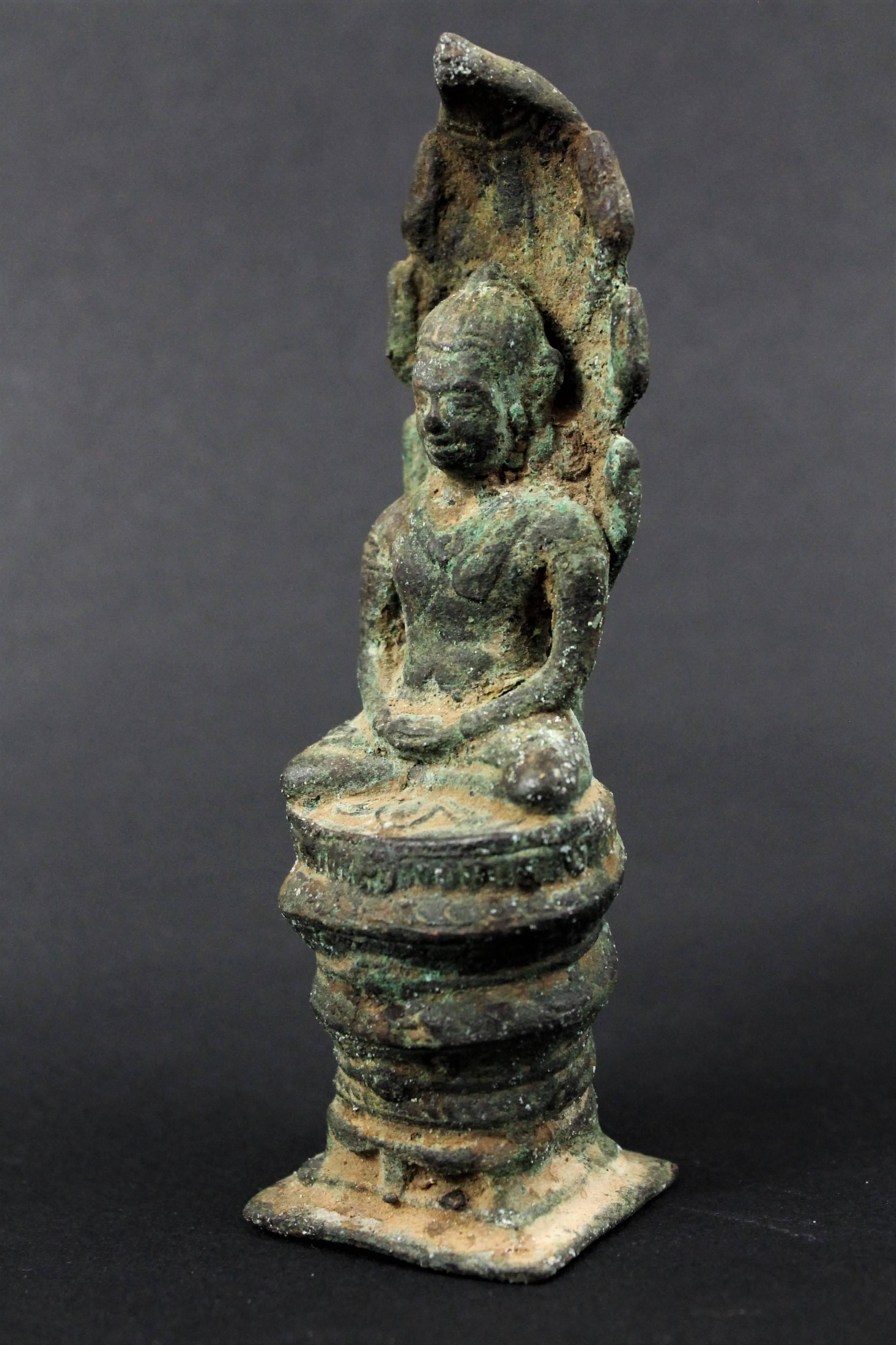 Bronze-Buddha im Mönchsgewand. Er sitzt auf dem gewundenen Naga, dessen Körper die drei Spiralen bildet. Die sieben Köpfe der Schlange bilden einen Baldachin, um Buddha während der Meditation zu schützen, mit einer Darstellung des Rades (Cakra) auf