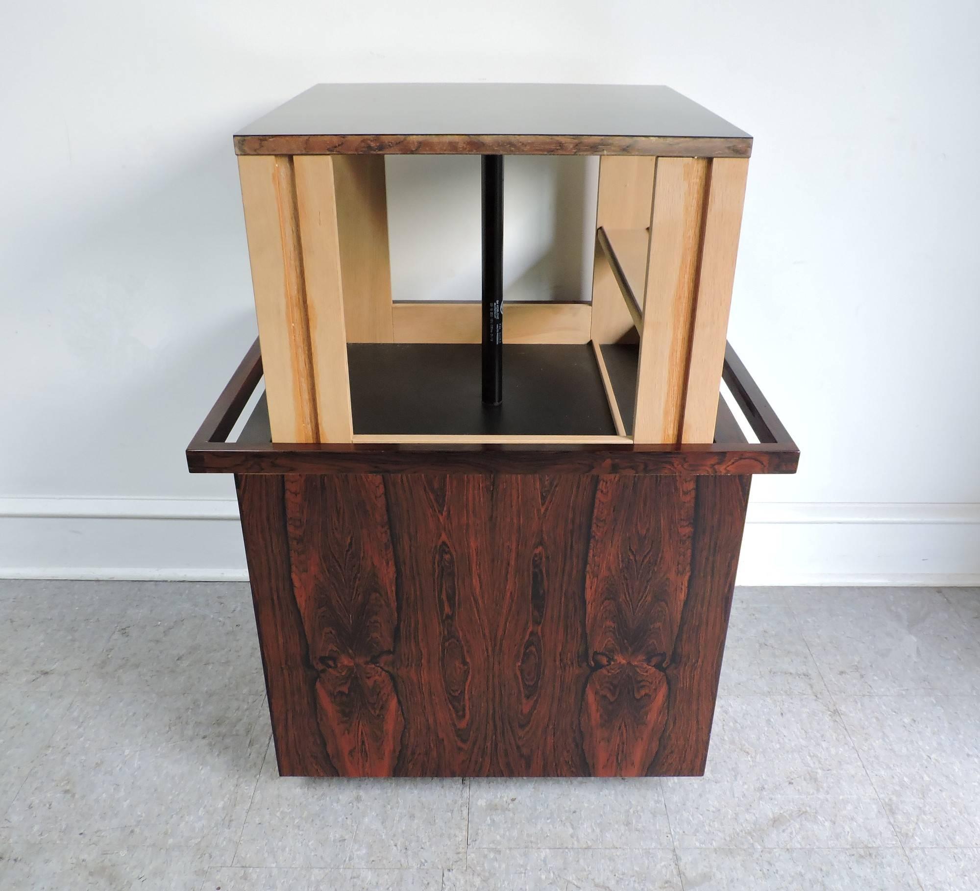 Very cool and handsome bar cart cabinet or table designed by Kai Kristiansen and made in Denmark in the 1960s. This cart has beautiful rosewood grain with a water resistant black formica top, and has a unique hidden storage compartment that rises up