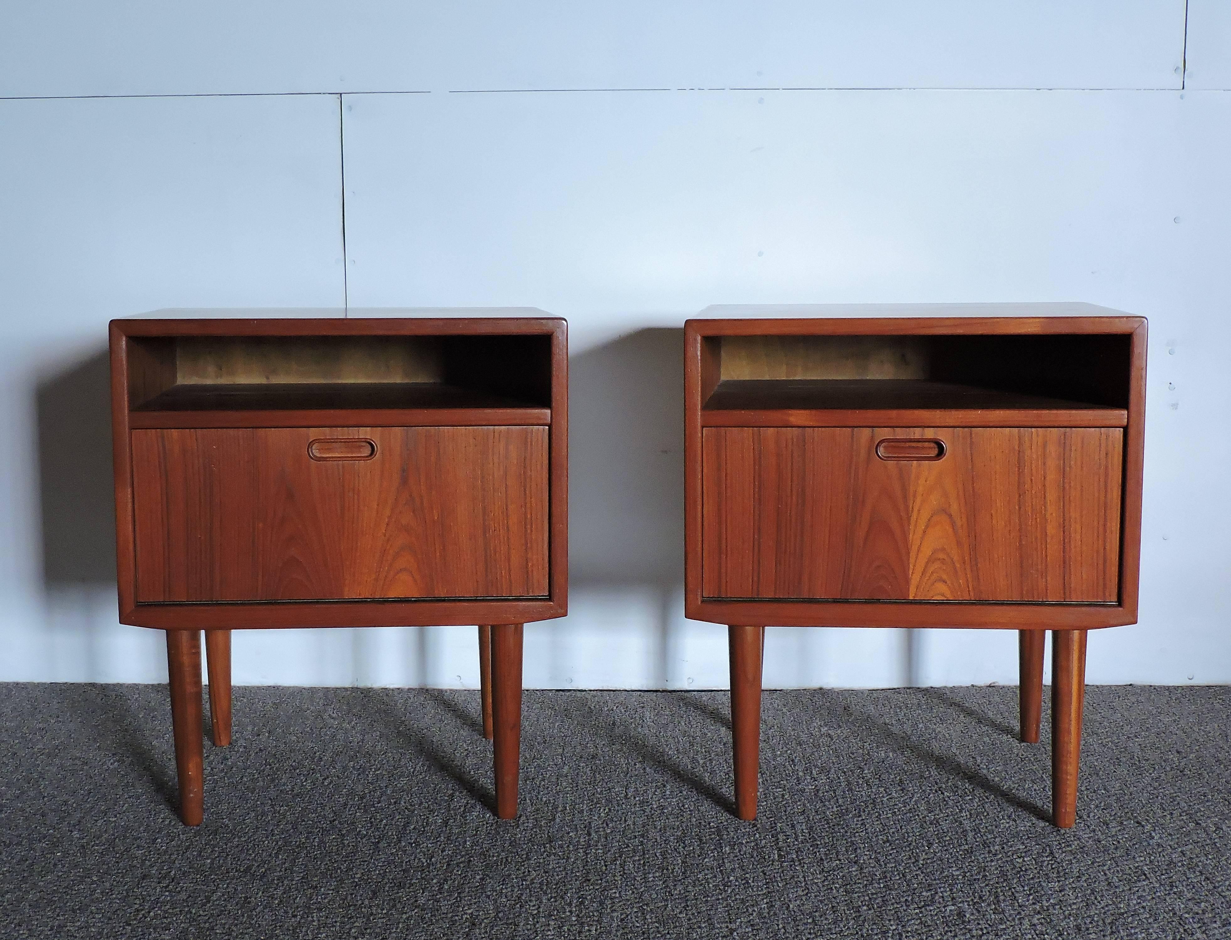 Wonderful pair of teak nightstands made in Denmark by Falster. These stands have an open shelf area, a drop-down door with a piano hinge, inset teak pulls, and tapered legs.
