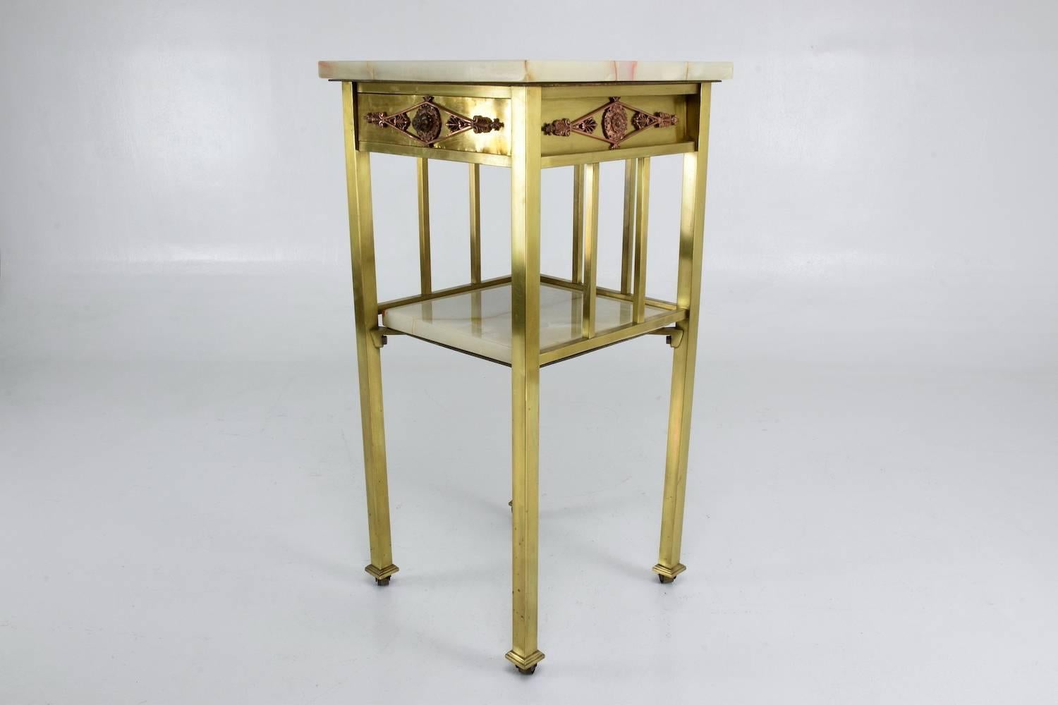 Beautiful 20th century French two-tier polished brass side table, nightstand or étagère with rollers, marble top, and shelf adorned with floral copper details around the upper drawer. 

---
We are an exhibition space and an online destination