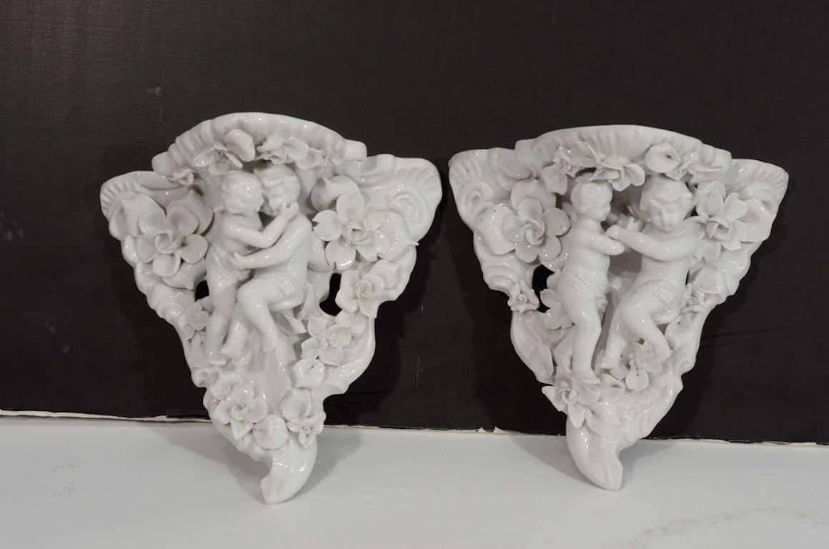 Two complementary white porcelain cherub wall shelves. They are marked only with a number; they seem Italian in origin but are otherwise unmarked. Each shelf depicts a pair of affectionate cherubs in different stages of embrace. The flowers