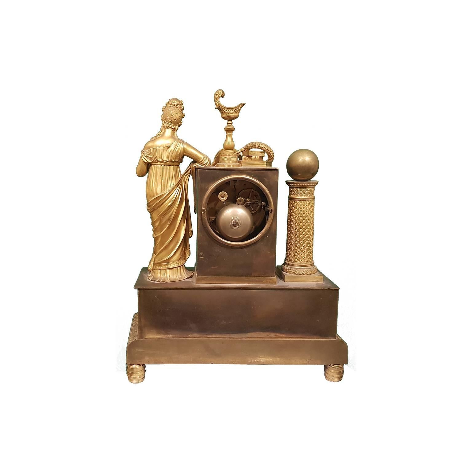 Antiguo reloj de estantería francés de bronce dorado rodeado por la Musa del conocimiento en el lado derecho y una columna con una esfera encima en el lado izquierdo. 

En el centro, el mecanismo se inserta en una decoración de forma rectangular