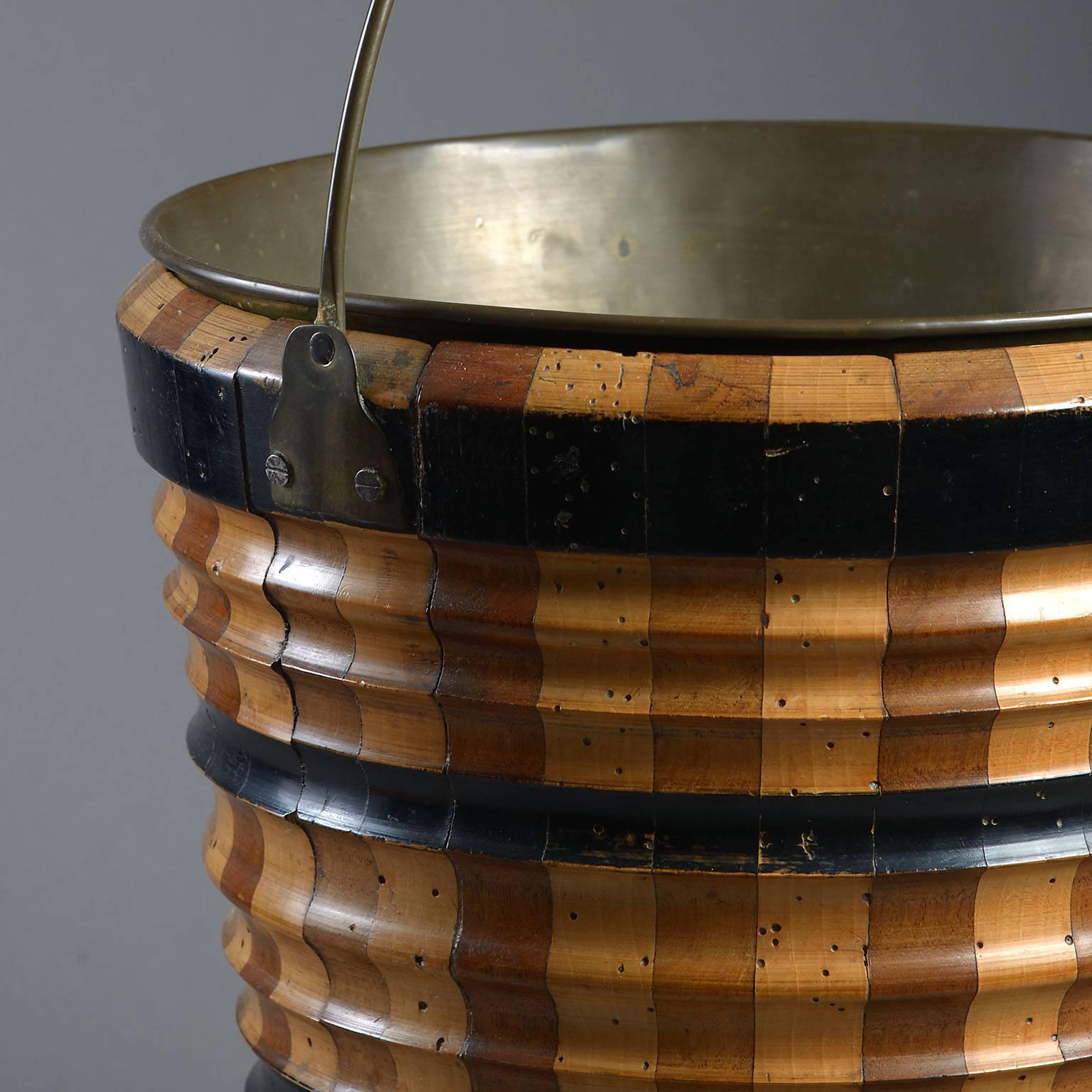 The lathe-turned body formed of contrasting staves of different timbers, coopered and highlighted with ebonized bands. This bucket is often referred to as a 'Teestoof' which was used for keeping a kettle warm over a bed of smouldering coals and