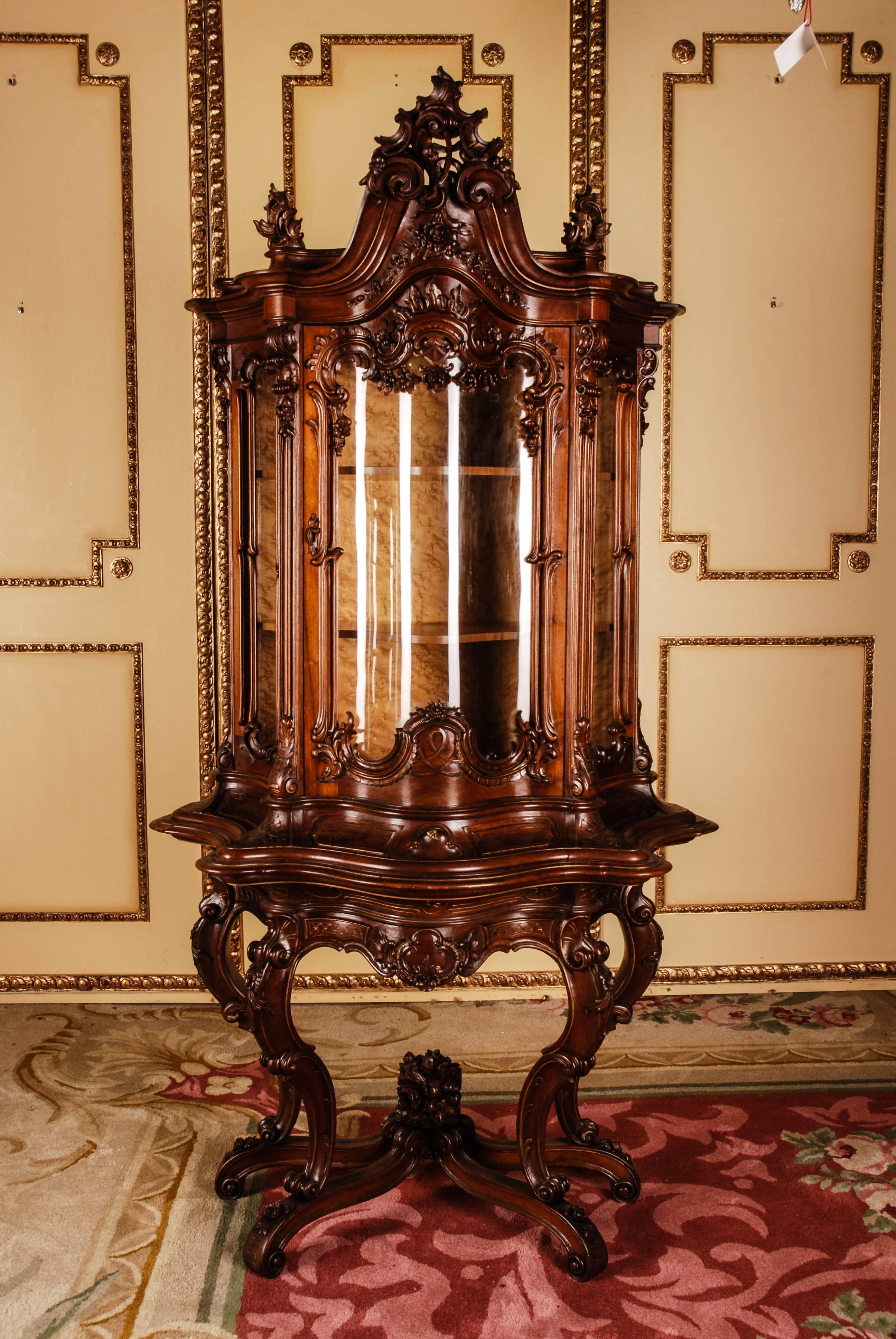 Massive walnut, finely carved and framed. High-rectangular, one-door, curved and three-glazed corpus on high slanting volute-like curled feet connected with crossed richly carved middle bridge. Three-sided scalloped, curved, incisive frame, rich
