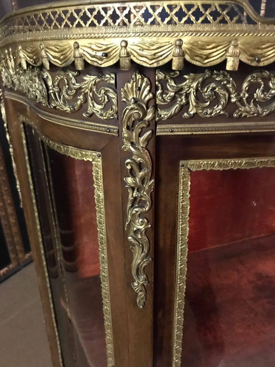 Rosewood on solid wood. High-aisle, one-door, curved and three-sided glazed body. On its curled feet, a draped frame.
Three-sided glazed display case. Slightly protruding top plate with brass gallery. Rich bronze fittings. Backboard covered with