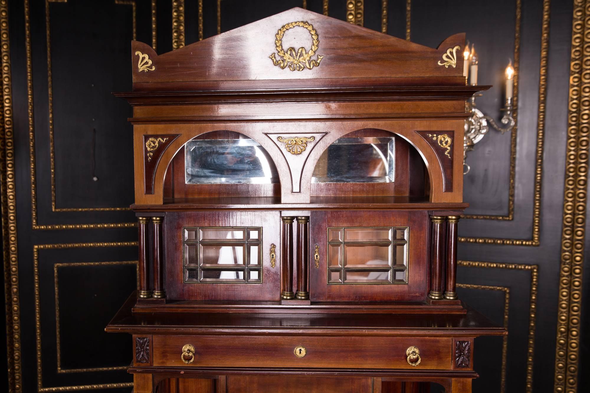 Mahogany on solid wood. High-arched, architecturally designed corpus, single-door base, open-sided shelf, flanked by columns with capitals and bases of finely chiselled bronze, slightly protruding top, including a wide drawer.

Excellent warm