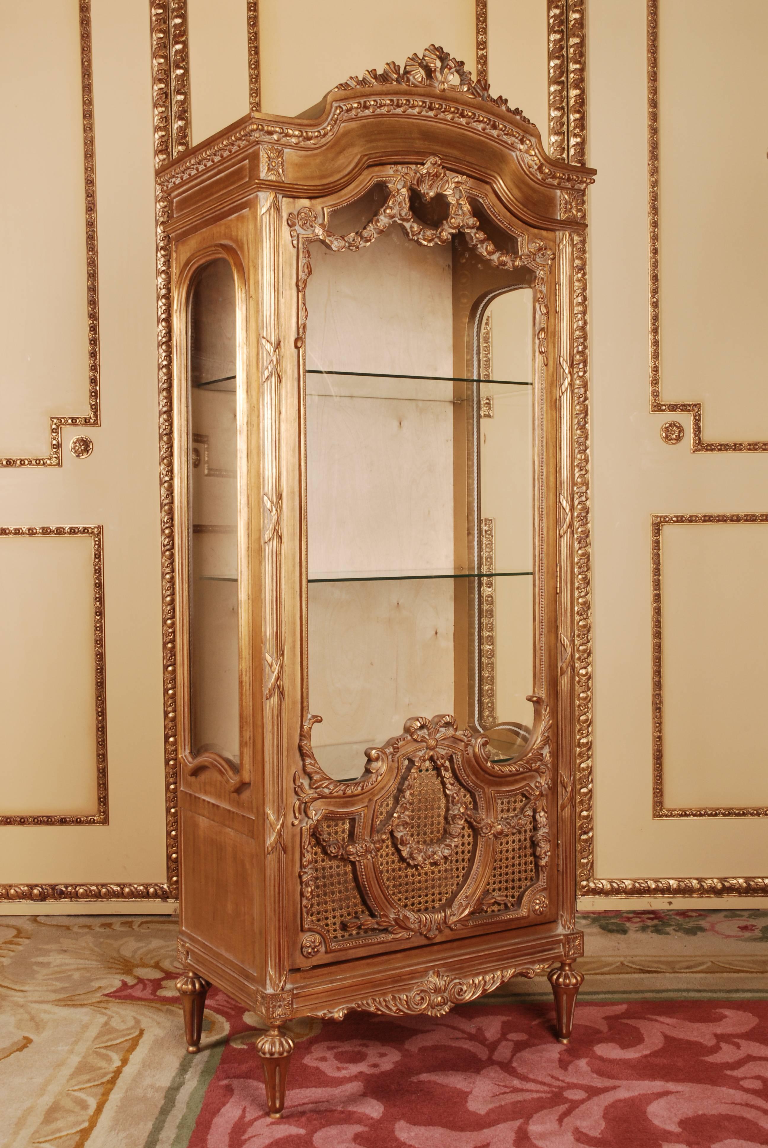 Solid beech, bole gilded. High-angled, three-sided, facet-cut glazed, one-door body on conical legs. The door is partially covered with wickerwork. The front, corners and sides are decorated with carved, classical elements such as garlands and
