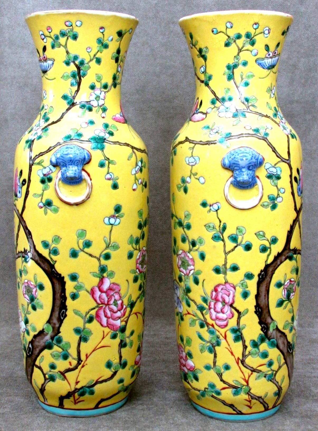 Both cylindrical yellow glazed bodies rising to waisted necks with flaring rims showing traces of gilt, decorated overall in enamels depicting leafy flowering branches, flower-heads and butterflies, sided by applied animal masks with rings.
(It is