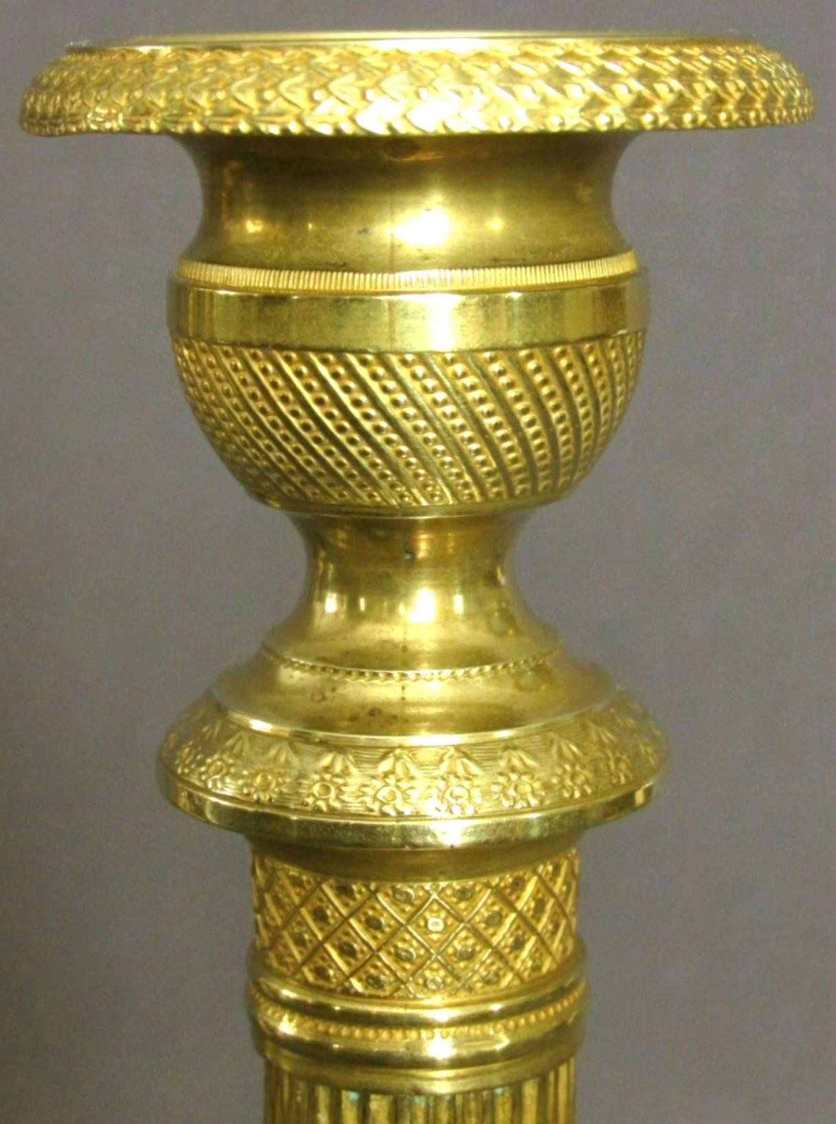 Both candlesticks showing reeded columns with finely milled capitals and urn-shaped nozzles with detachable drip pans, rising from circular bases decorated with milled geometric motifs. The underside of each base bearing faintly stencilled inventory
