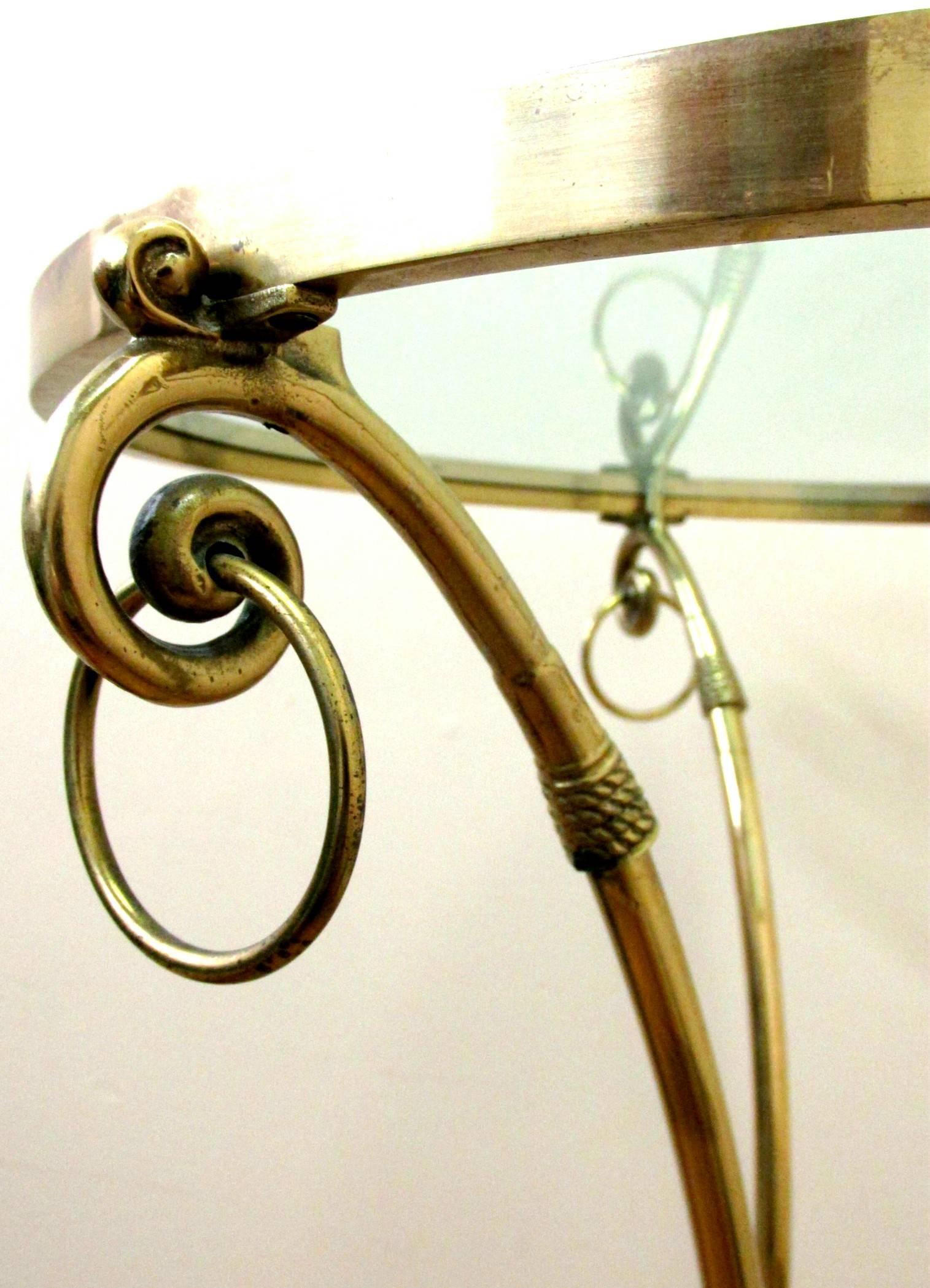The brass frame showing three slender supports rising from hoof feet, braced by a circular stretcher surmounted by a circular glass top, the brass frieze decorated with three scrolled capitals with ringed elements.

International clients are tax
