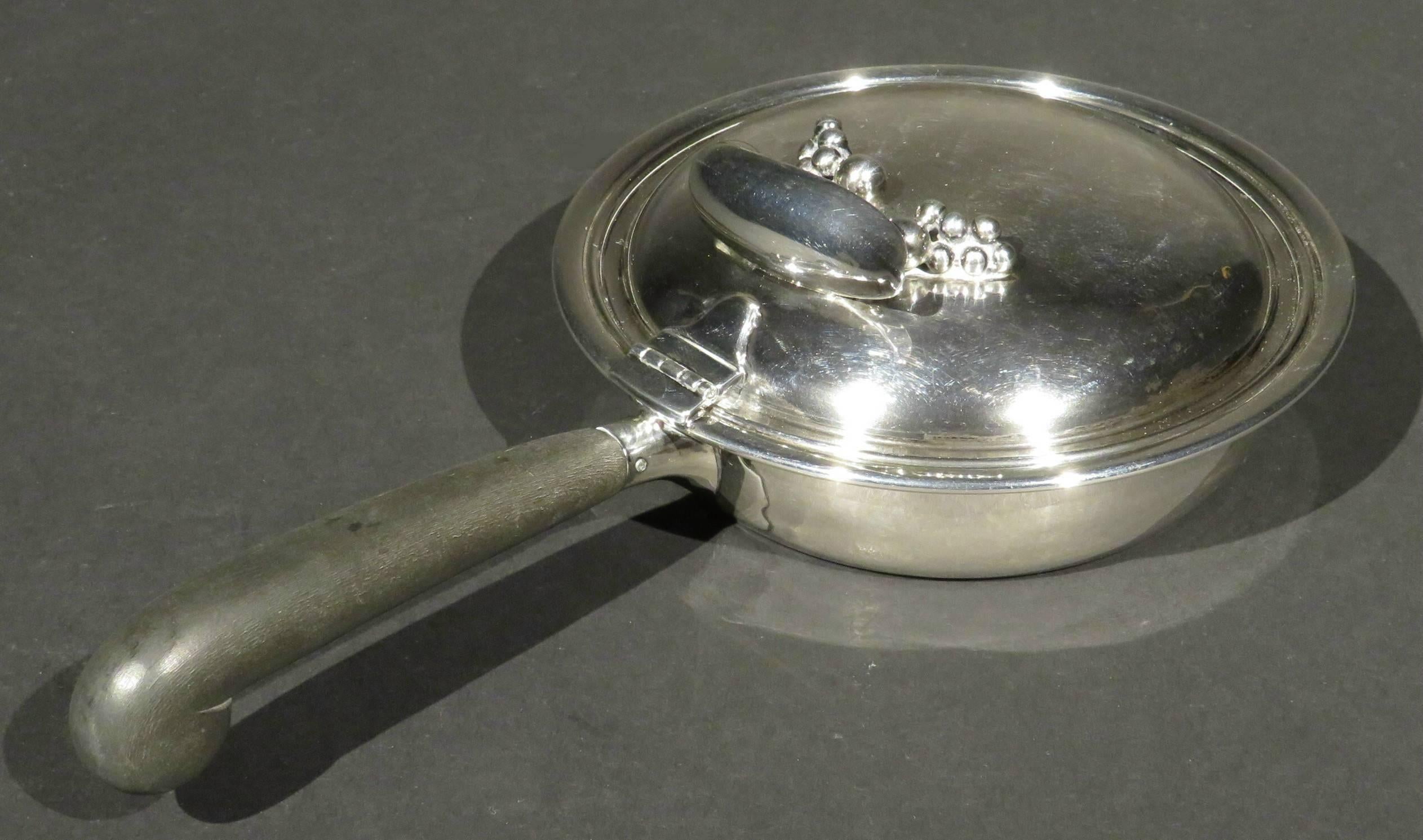 The sterling silver body fitted with an ebonized wood handle, affixed with a hinged lid having a subtle planished surface and an applied organic inspired element. 
Carl Poul Petersen (b. Denmark 1895 – d. Canada 1977) apprenticed with Georg Jensen