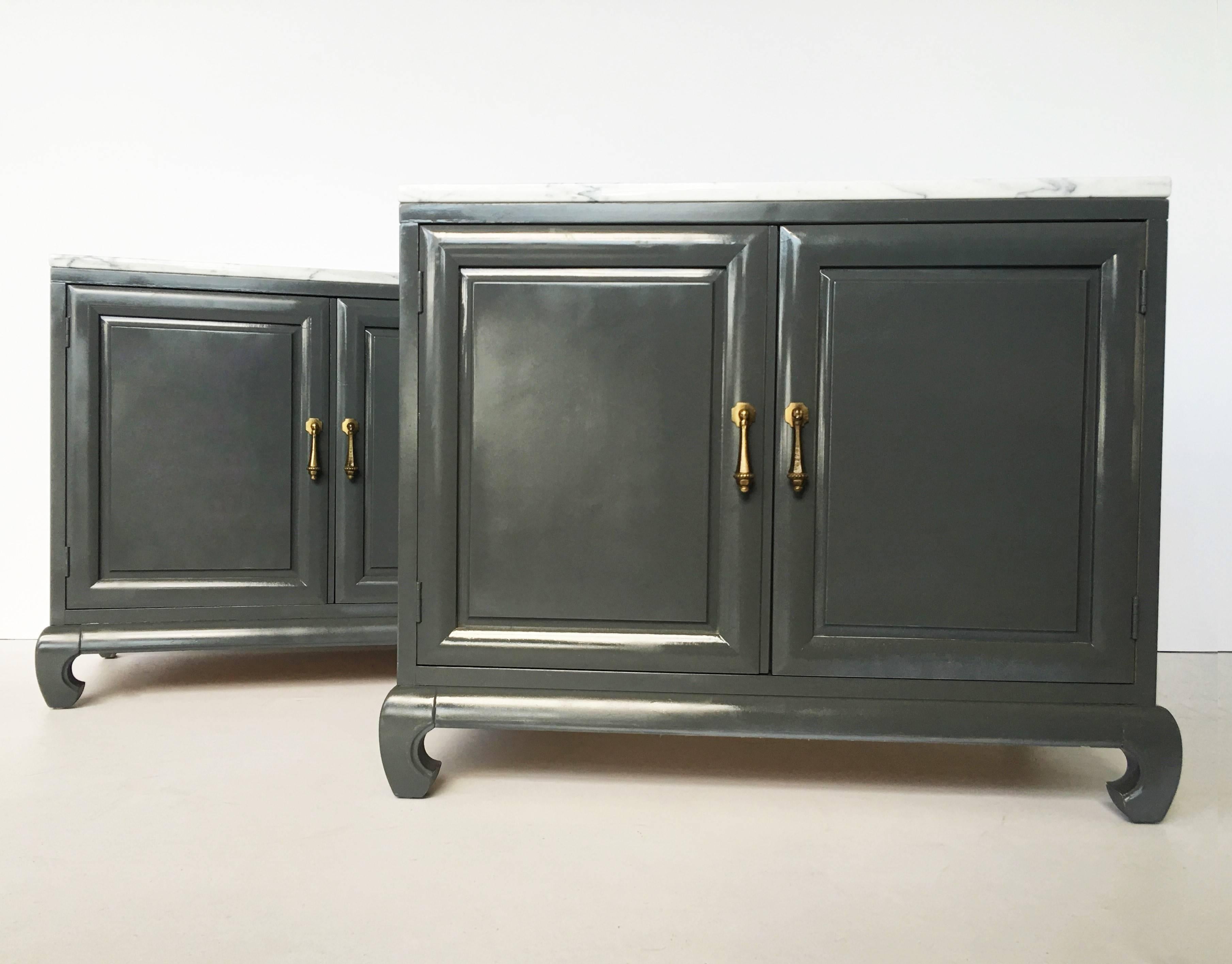 A pair of stunning Asian influenced styling cabinets made of wood and lacquered in a dark silver-gray tone. Each cabinet has two doors in the front with brass hardware pulls. Ming style turned legs. The interior has one shelf. These are beautiful