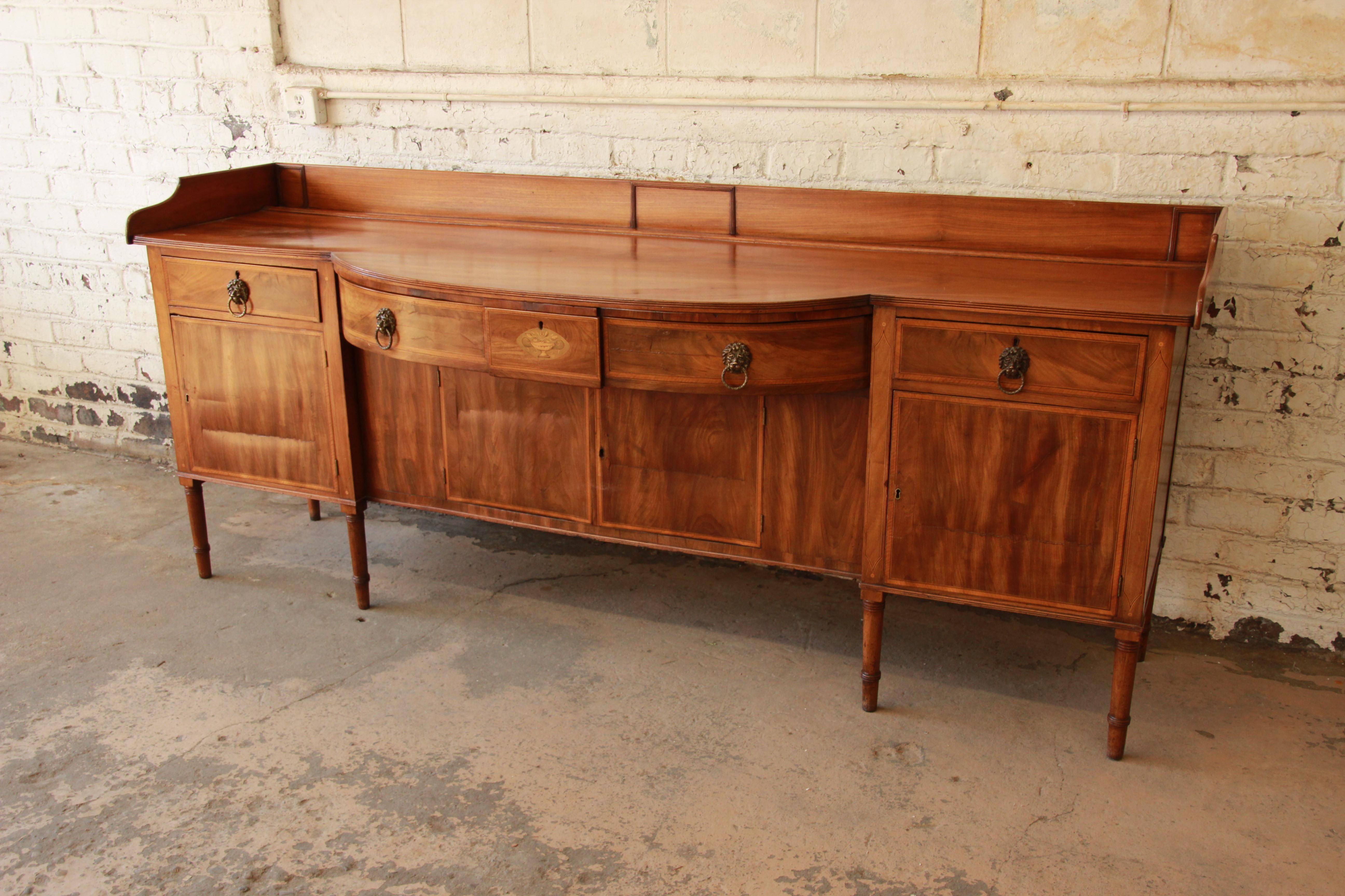 An outstanding inlaid mahogany English sideboard, circa 1820. This monumental piece features stunning inlaid details, gorgeous flame mahogany wood grain, and original lion's head brass drawer pulls. The sideboard offers ample room for storage, with