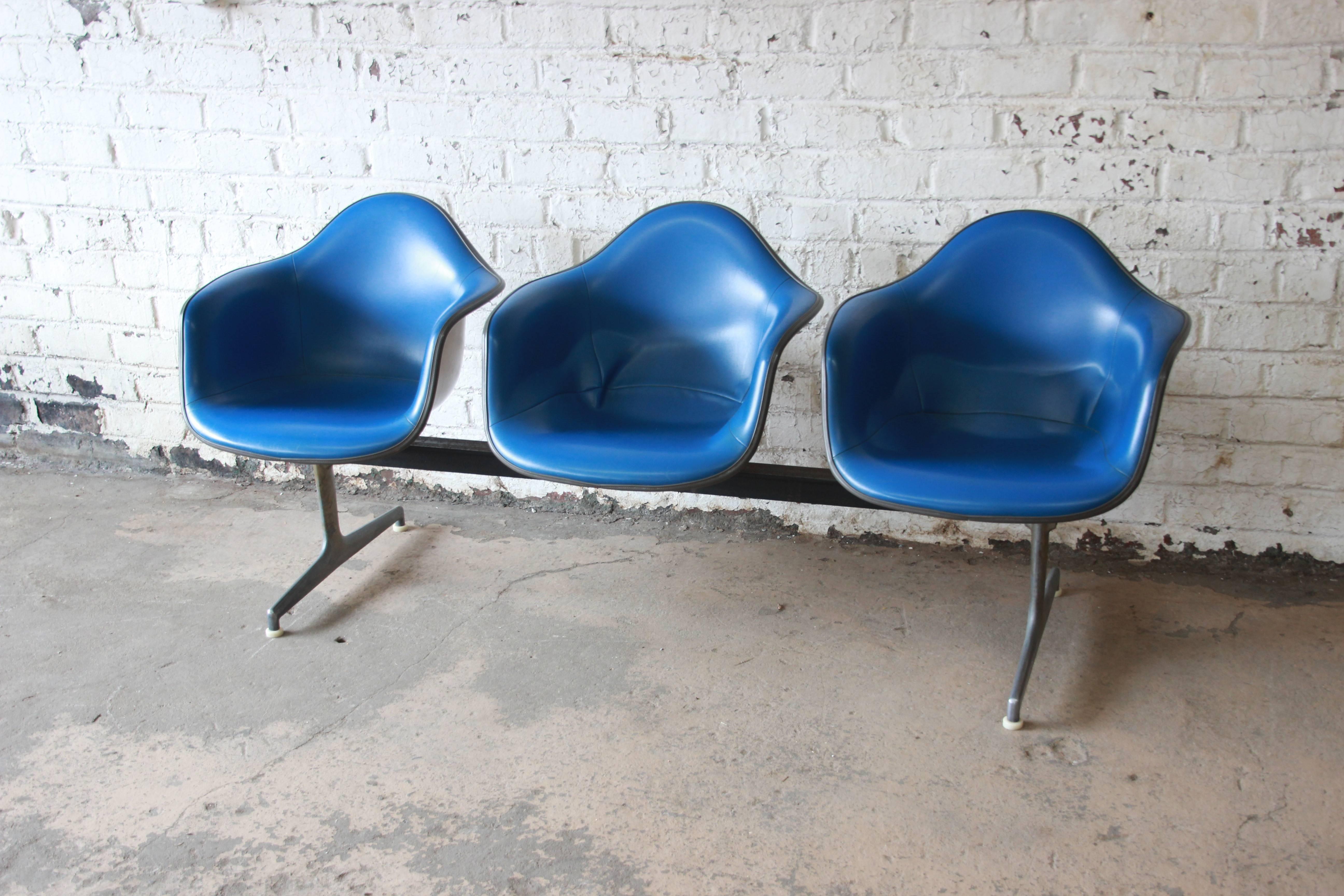 Three tandem shell chairs in original blue leather designed by Charles & Ray Eames in the 1960s. The chairs sit on a metal and aluminium frame with three iconic shells chairs attached. These items were originally seen in airports and offices and now