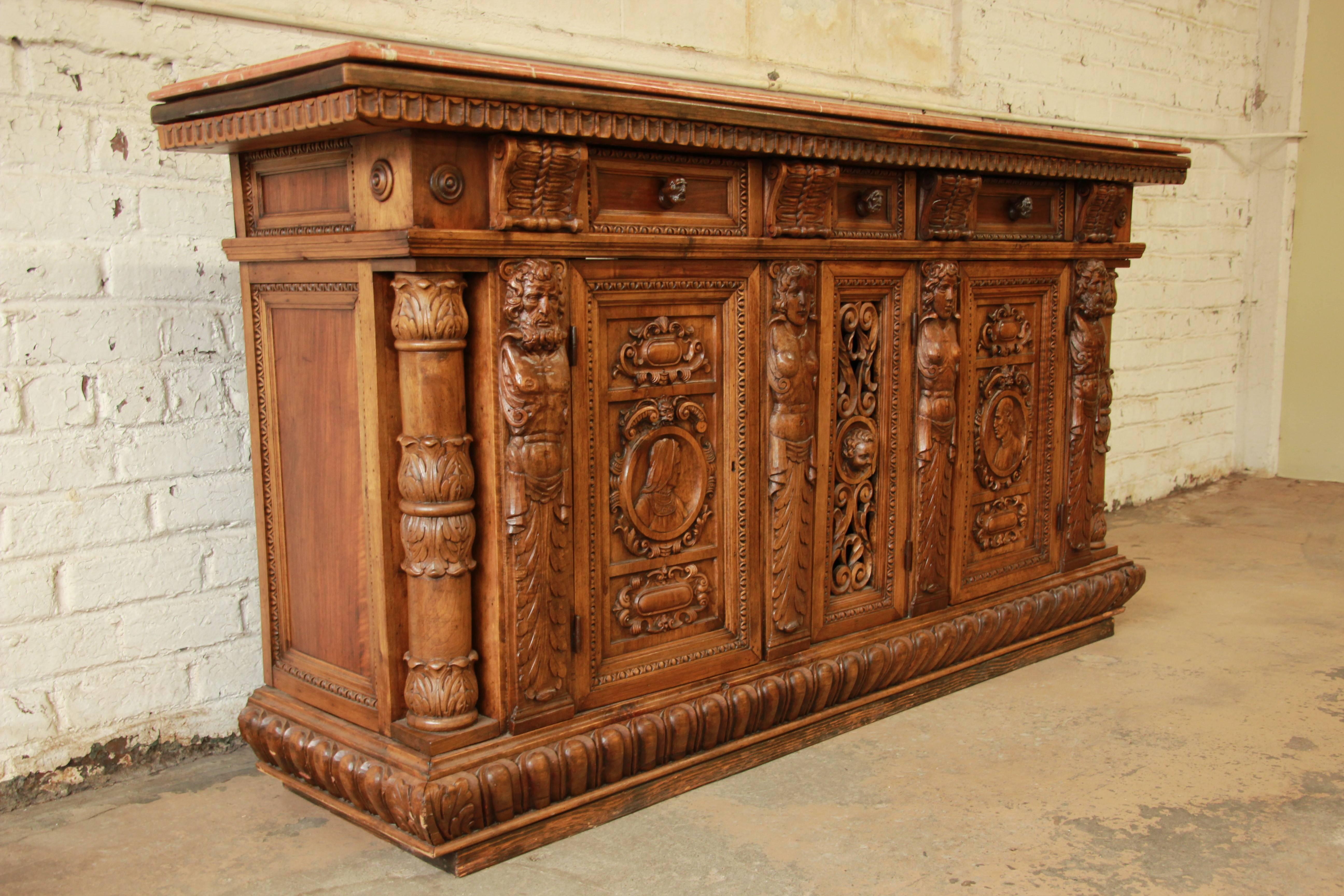 Offering a stunning ornate hand-carved 19th century Belgian sideboard buffet bar cabinet with marble top. The cabinet features stunning carved wood details, including twelve carved faces and carved columns on each end. It is made from mixed wood -