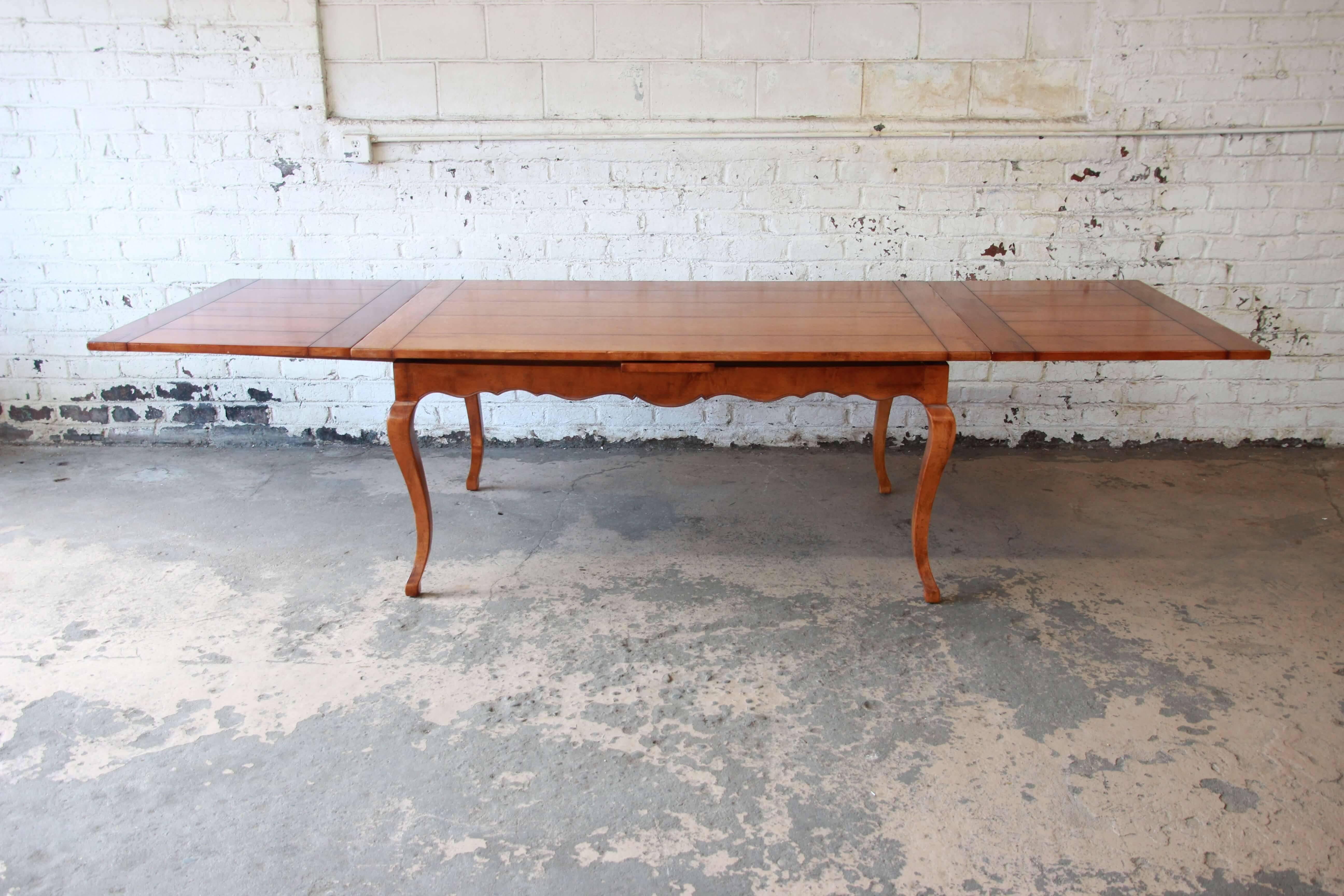Offering a beautiful vintage solid maple extension dining table by Baker Furniture's Milling Road division. The table features a designed distressed look with French legs and apron. The two leaves are a refectory leaves and are stored underneath the