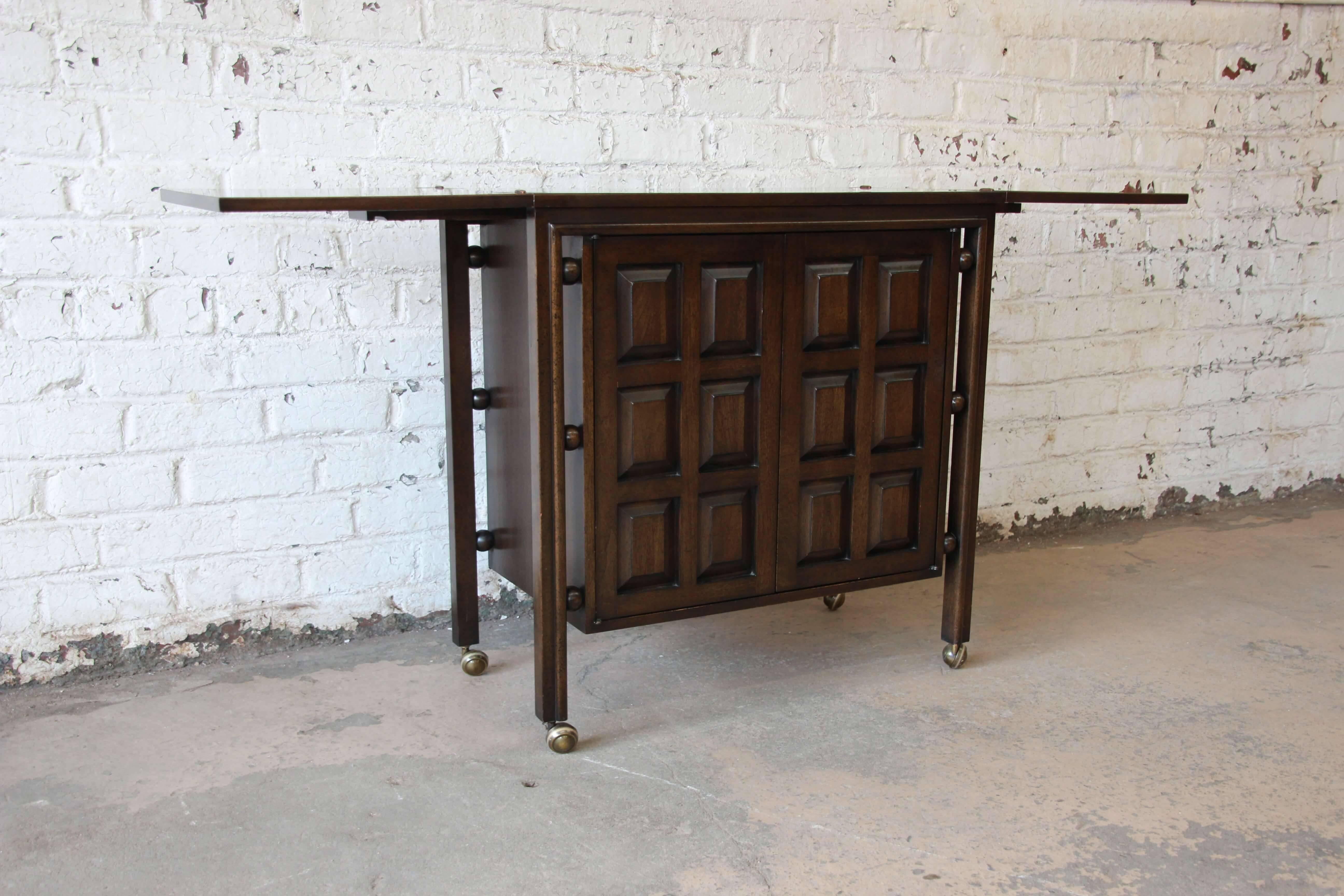 Offering a gorgeous vintage dark walnut Spanish Colonial style bar cart by John Widdicomb. The cart features rich walnut grain and a unique Spanish design. The doors open to reveal a single dovetailed drawer, three glass shelves, and wine glass