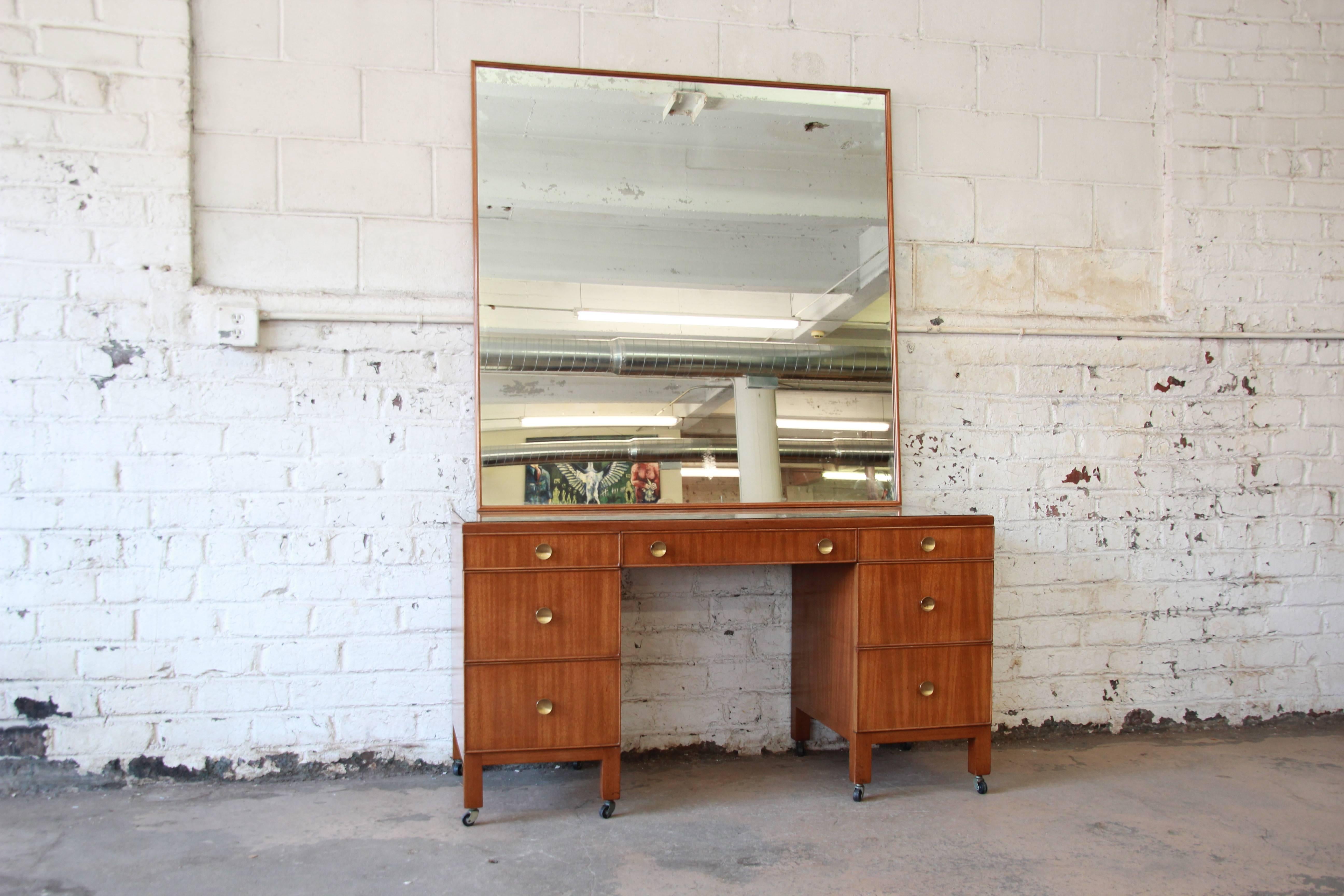 Offering a very rare and early vanity and mirror by Edward Wormley for Dunbar. The vanity offers seven drawers for ample storage. It has original brass pulls with an inset glass top and sits on casters. The large matching mirror extends to nearly