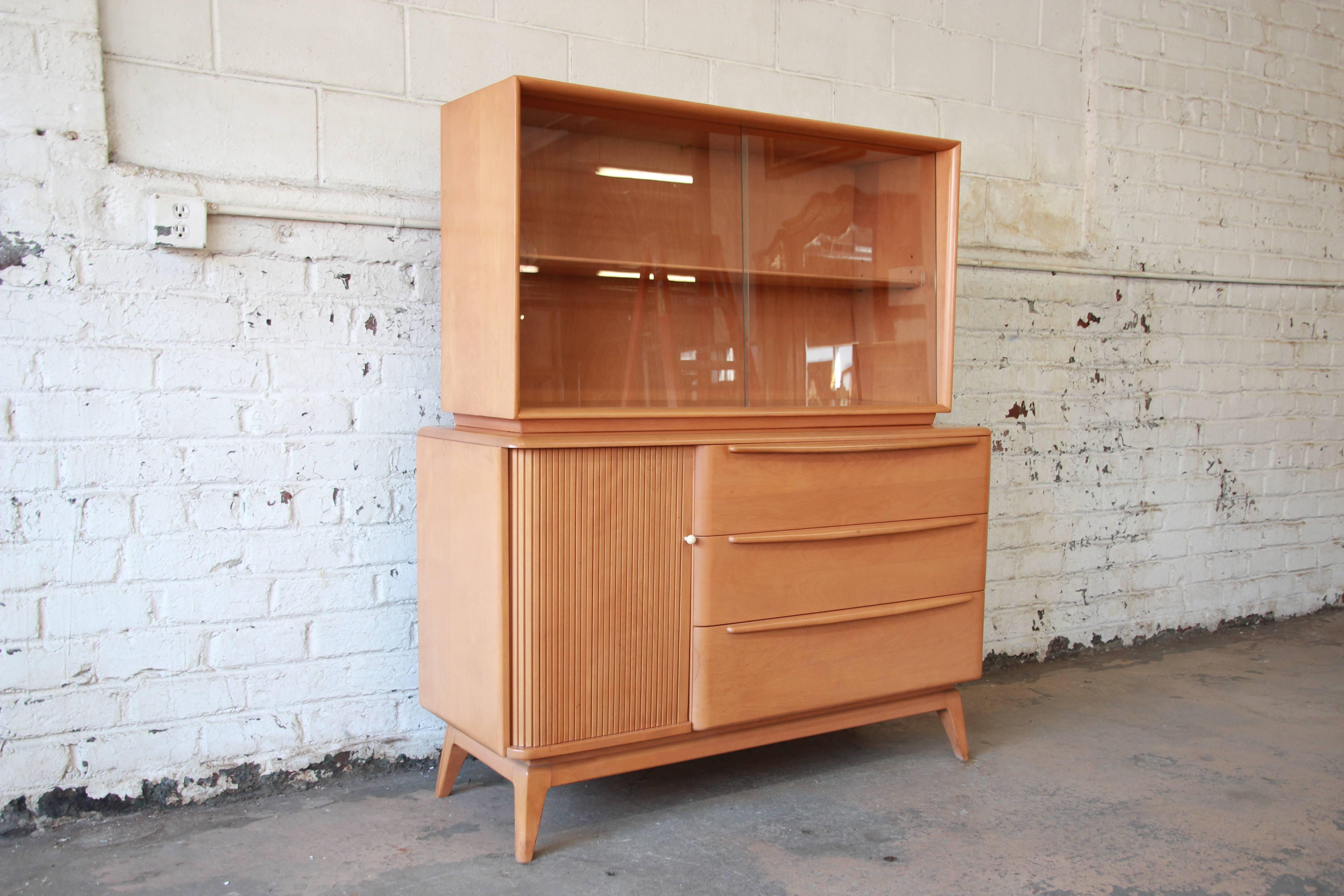 Offering a very nice Heywood-Wakefield sideboard with glass-front hutch top. The piece has a tambour door on the left side of the sideboard and three large drawers on the right. The removable top hutch has nice glass sliding doors and an adjustable