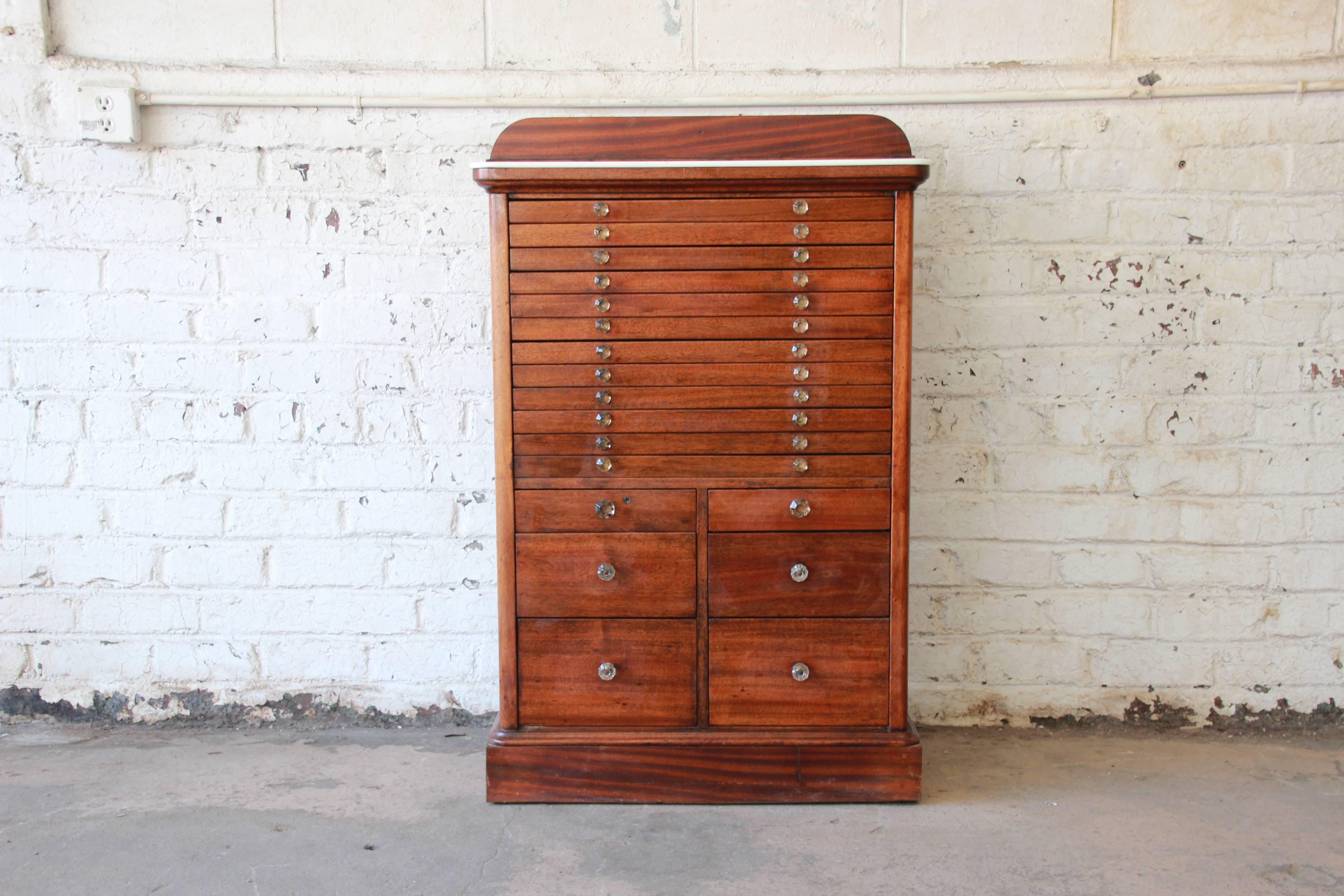 Offering a rare and unique antique mahogany dental cabinet. The cabinet features sixteen graduated drawers, with glass drawer pulls and metal interiors. It has a nice milk glass top and a backsplash. Considering age and use, the cabinet is in very