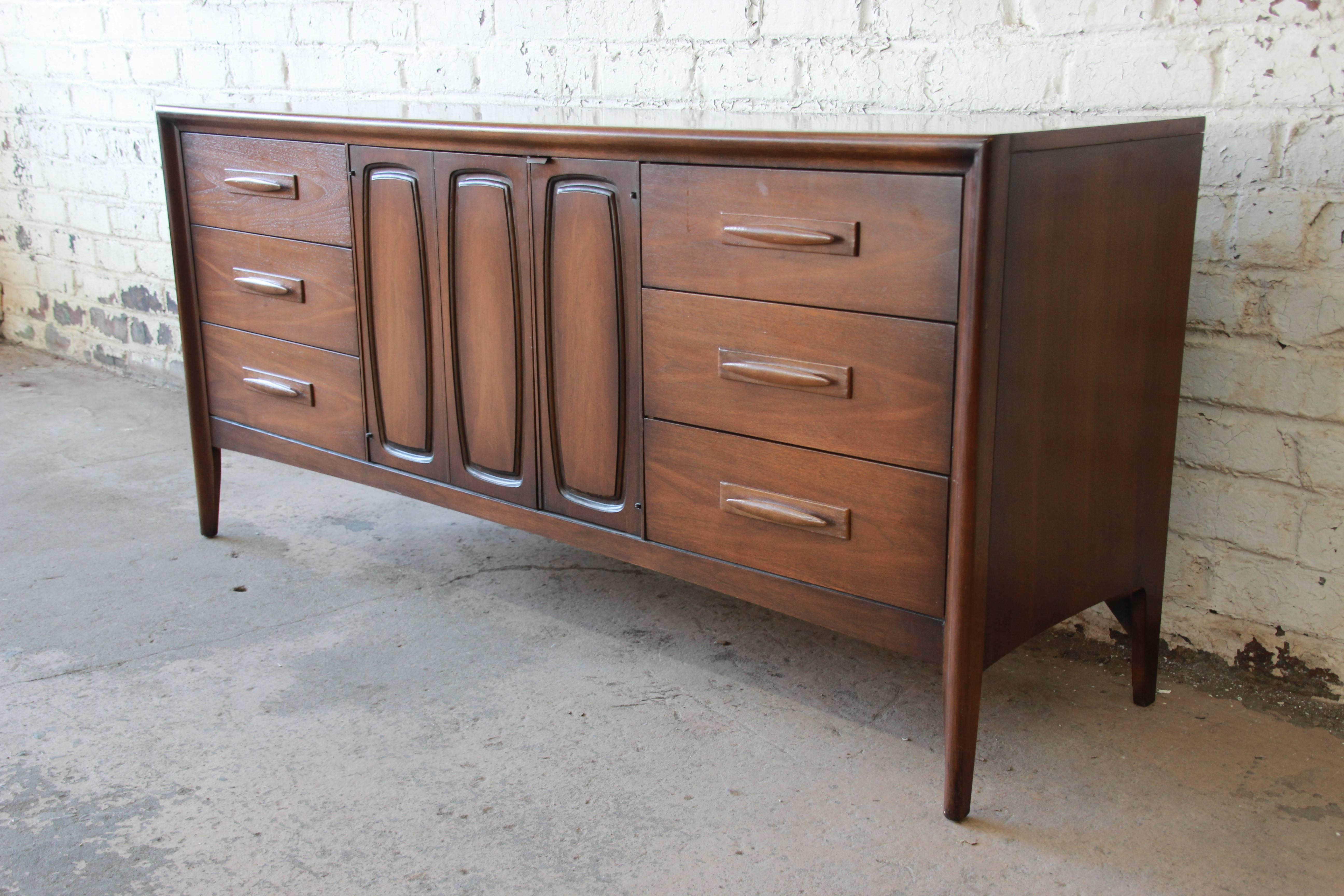 A very nice Broyhill Emphasis Mid-Century Modern sculpted walnut credenza or triple dresser. The credenza has clean midcentury lines, beautiful walnut wood grain, and nice sculpted walnut drawer pulls. It offers ample room for storage, with nine