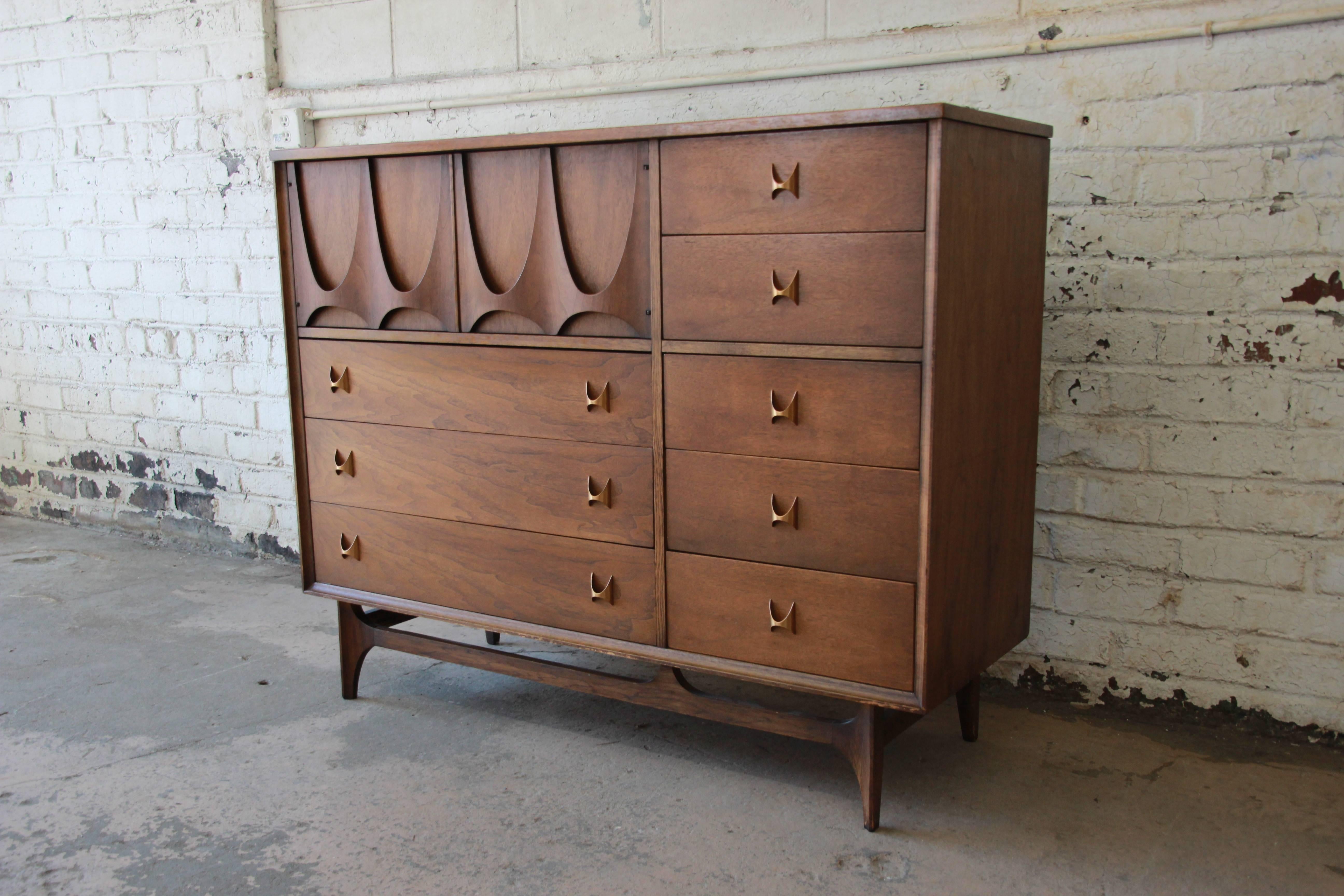 An outstanding Mid-Century walnut highboy dresser or chest of drawers from the Broyhill Brasilia line, circa 1960s. The chest features eight dovetailed drawers with signature brass handles. Includes open storage with two dividers behind sculpted