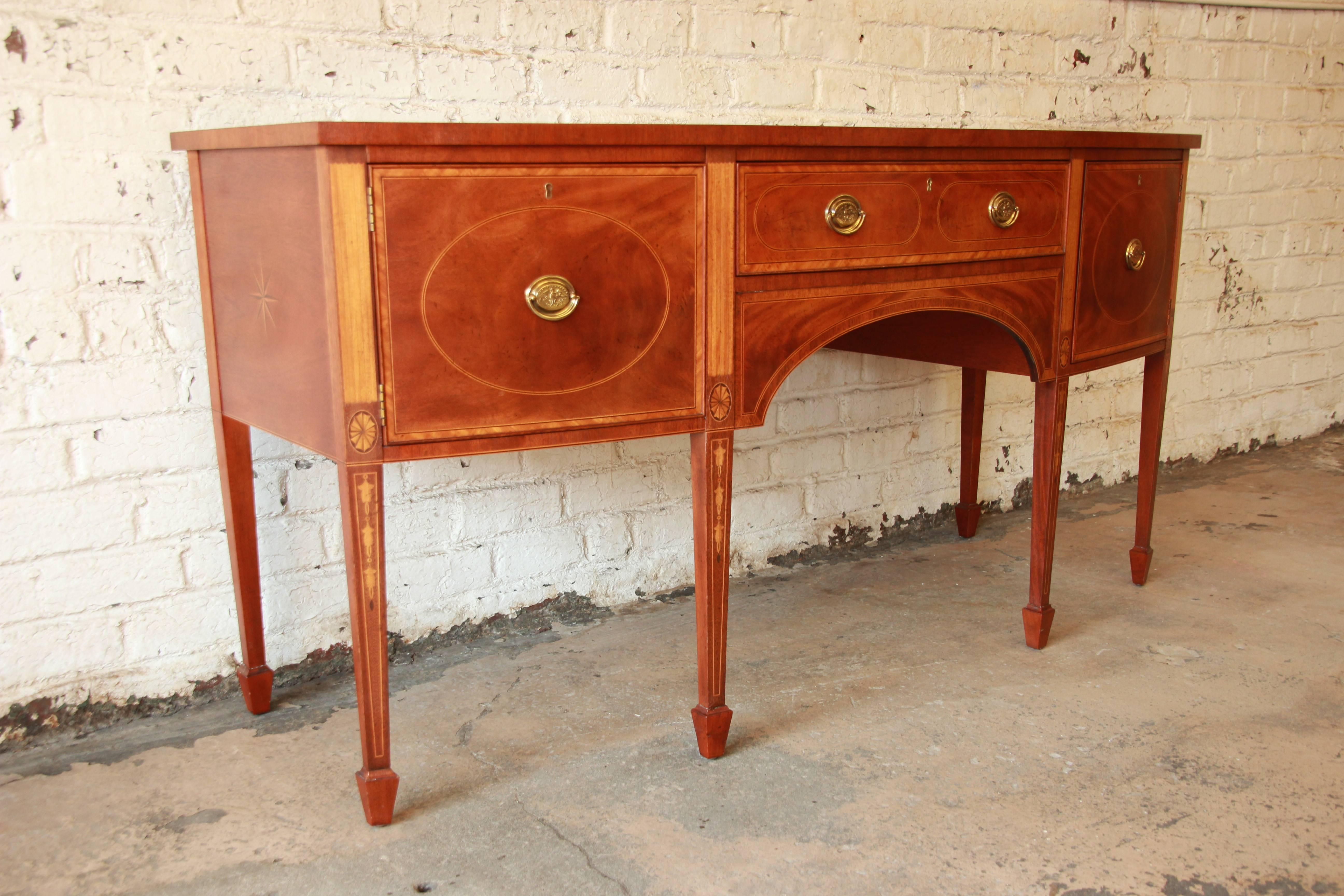 A stunning inlaid mahogany American sideboard buffet from the Historic Williamsburg line by Baker Furniture. This sideboard features intricate inlaid details and brass pulls with eagle design. The top middle drawer is felt-lined for silverware. The