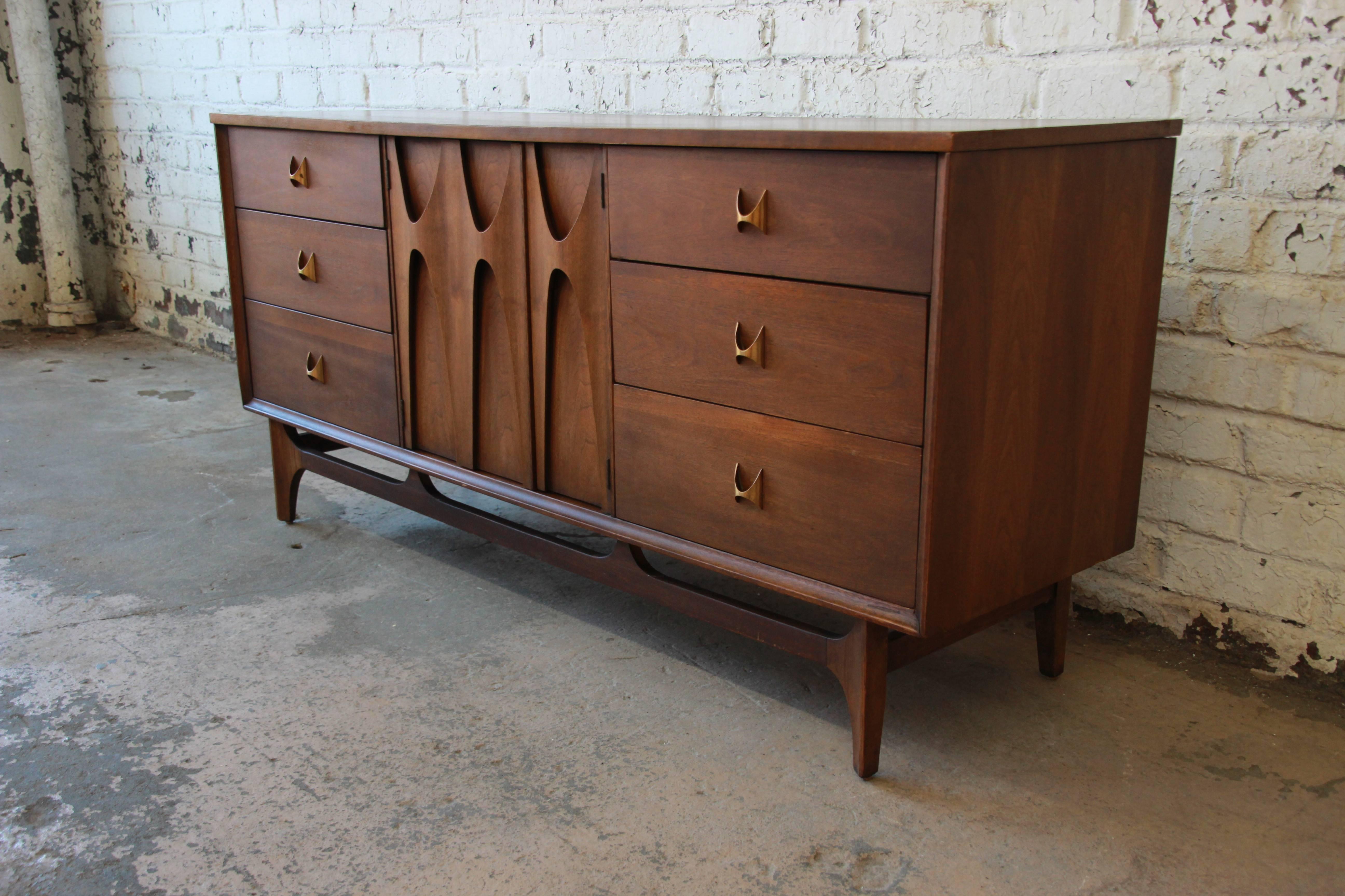 An iconic Broyhill Brasilia Mid-Century Modern sculpted walnut nine-drawer dresser. The dresser features gorgeous walnut wood grain, with sculpted arches and original pulls. The centre sculpted cabinet doors open up to three drawers with an