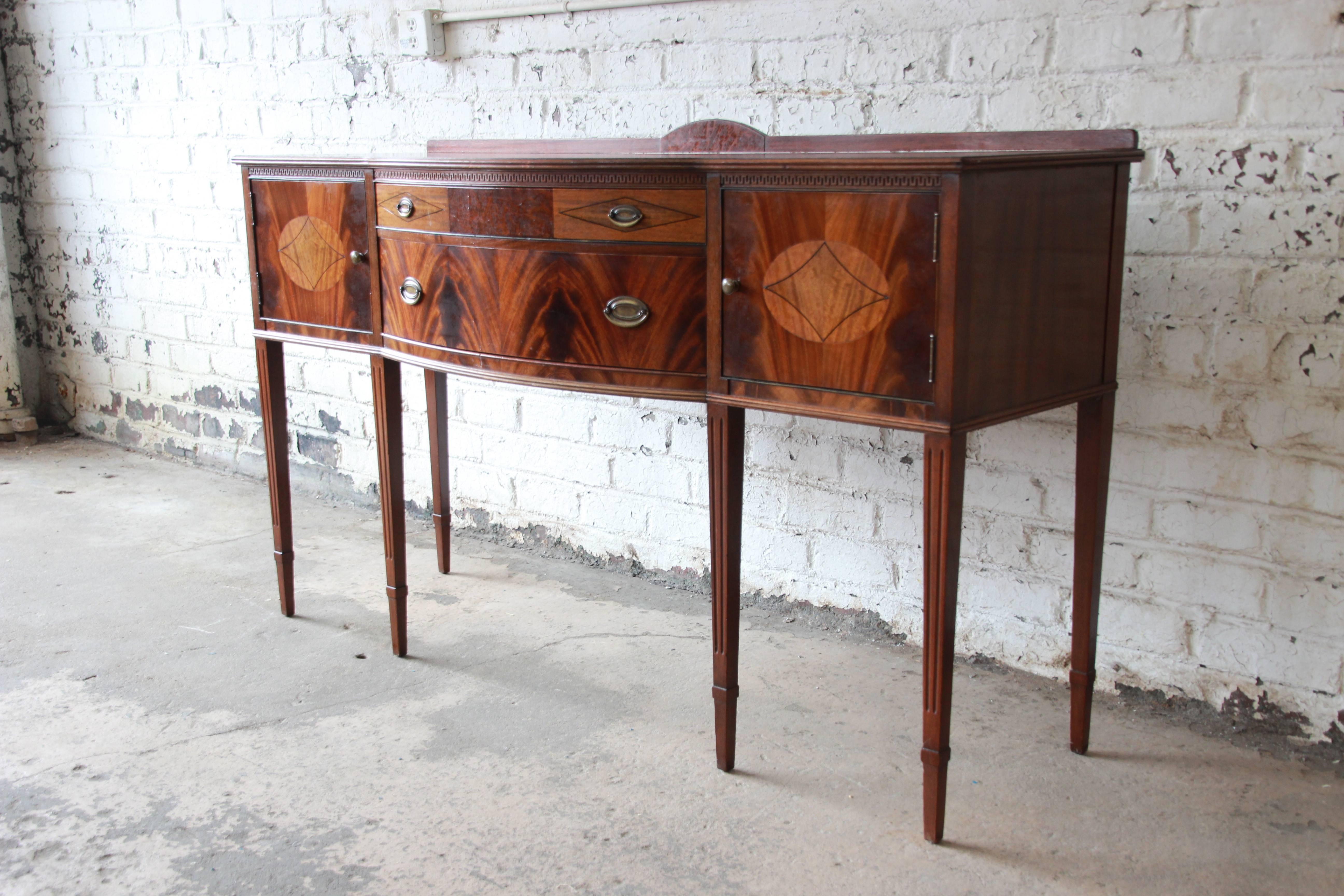 An outstanding Hepplewhite style inlaid flame mahogany sideboard buffet by Limbert. The sideboard features stunning flame mahogany wood grain with gorgeous inlaid details. It offers ample room for storage, with two central drawers and two more