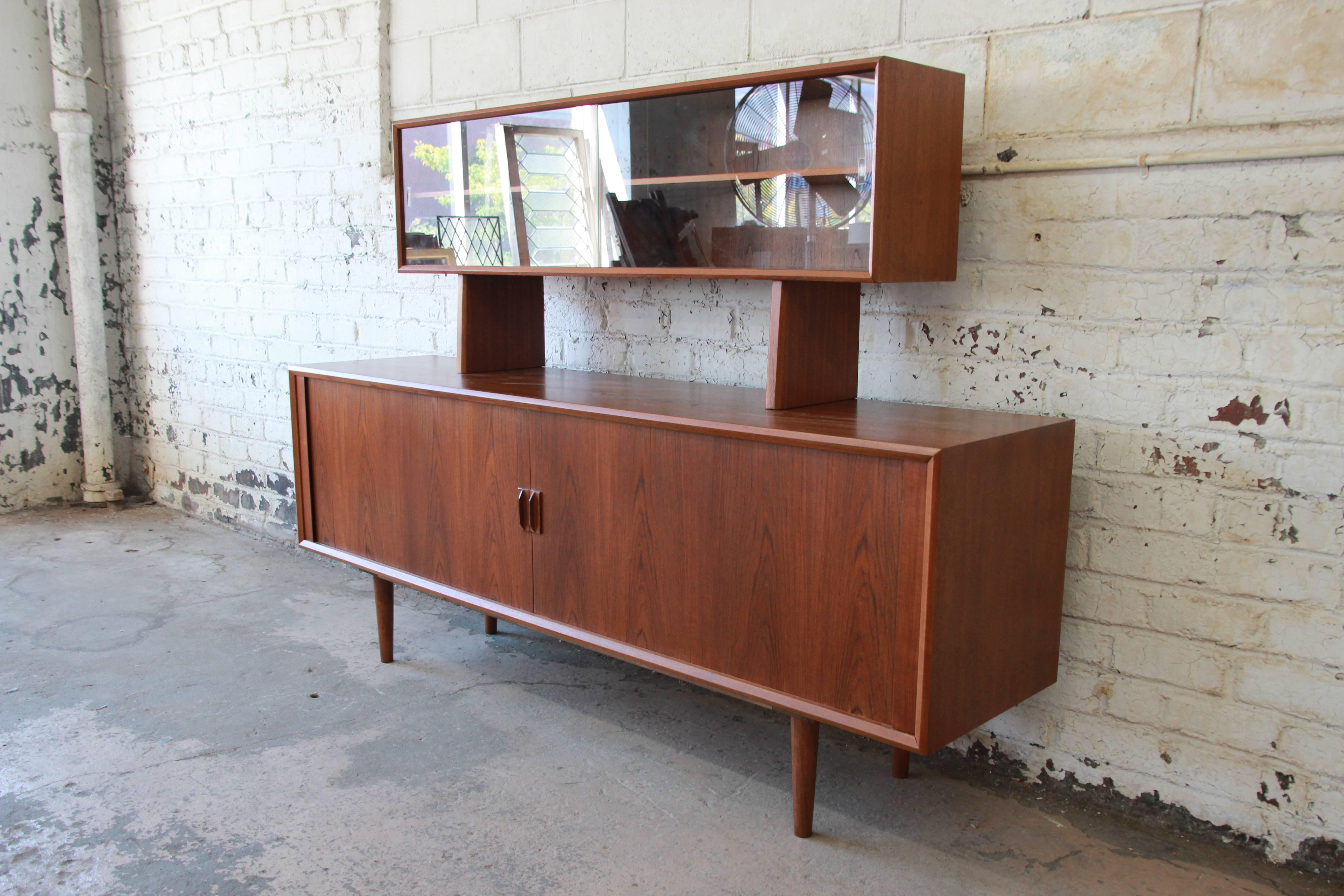 An outstanding Danish Modern teak tambour door long credenza with glass front hutch top designed by Ib Kofod-Larsen for Faarup Møbelfabrik. The credenza features stunning teak wood grain and sleek Danish design. It offers ample room for storage,