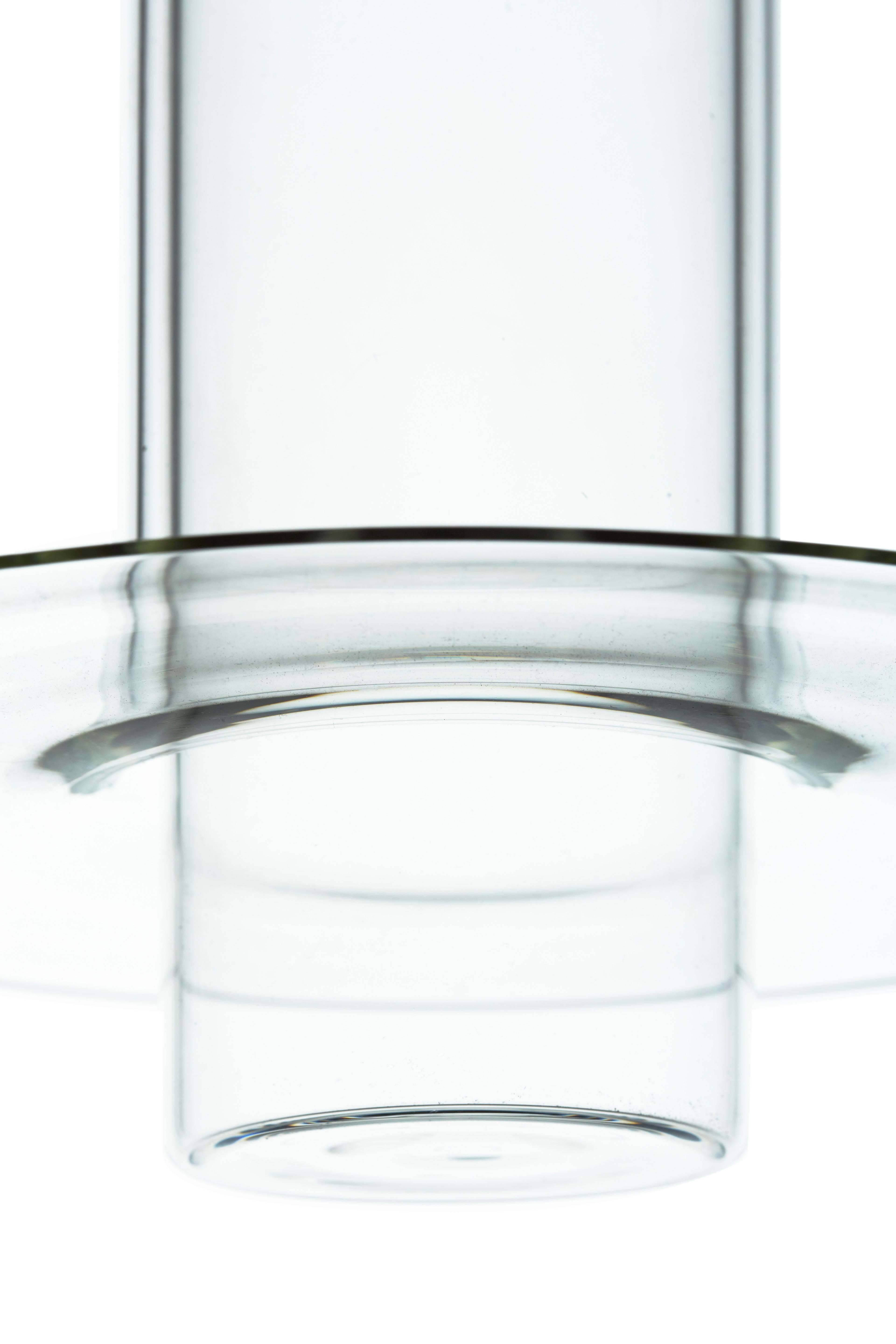 Pawsons Entwurf setzt einen handgefertigten Glaszylinder in einen anderen, wobei sich der äußere Zylinder an seinem unteren Rand zu einer raffinierten Scheibenlippe erweitert. Die Lampe wirft ihr Licht nach unten, aber ihr klarer Körper leuchtet
