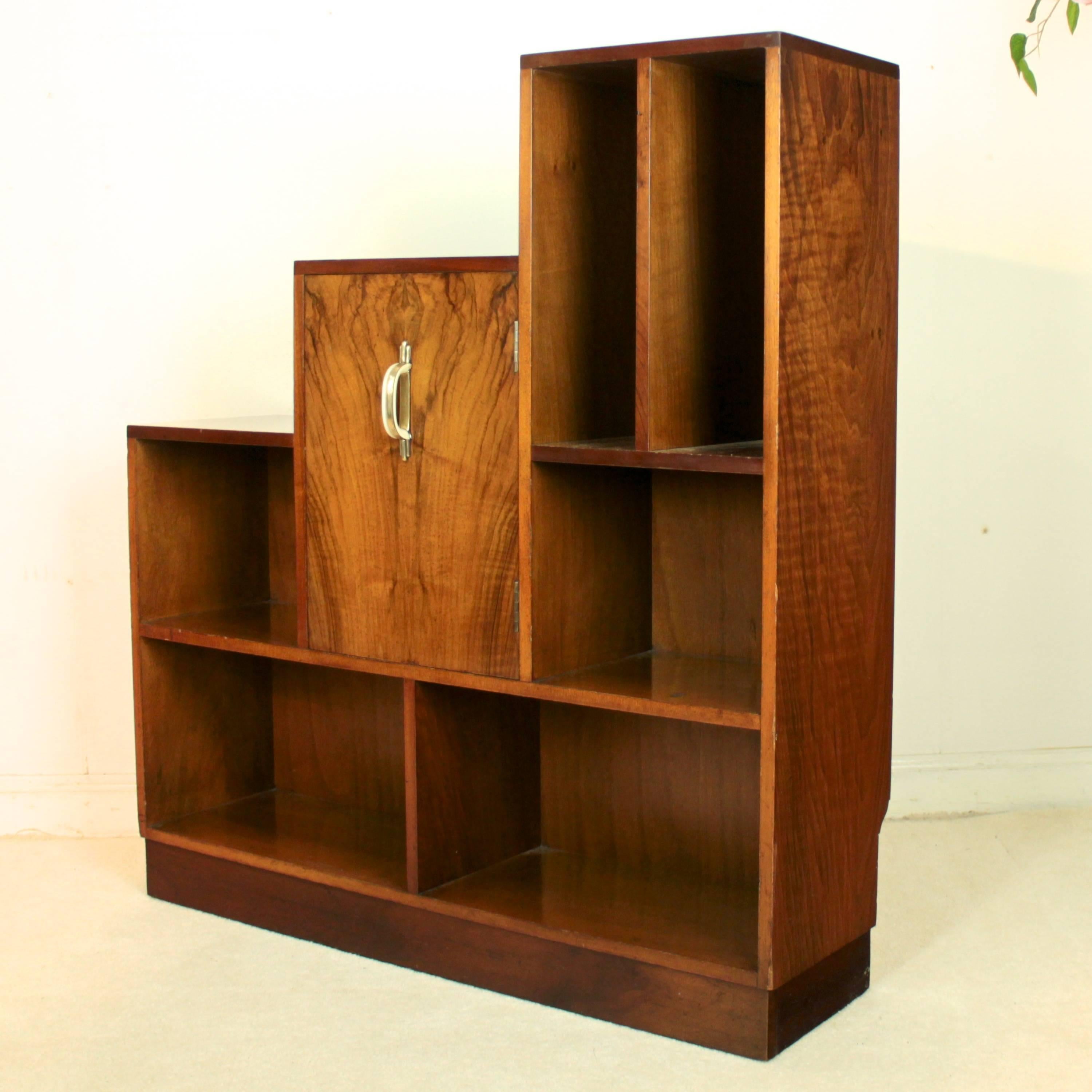 A stylish Art Deco walnut open bookcase made in 1936 by the famous British furniture makers E Gomme. Measuring just over 36in wide it has a stepped top with various open shelves and a central cupboard door with original cream bakelite and brass