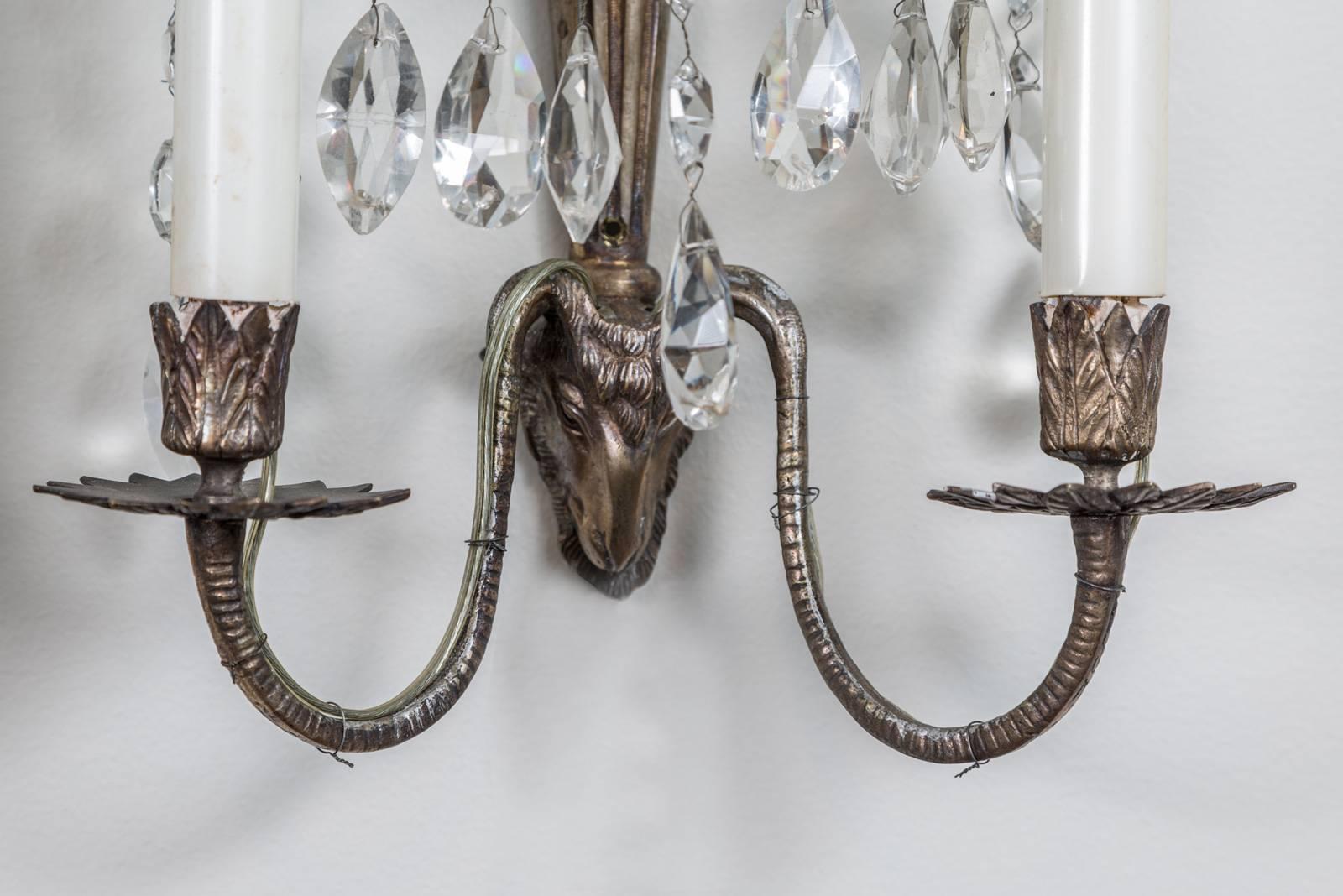 Two pairs of elegant Swedish crystal and metal two-arm sconces, each centered by a ram’s head from which issues a vase form with eleven sprays of beaded crystals, above that is a tall crystal faceted spire from which emanates five sprays of beaded