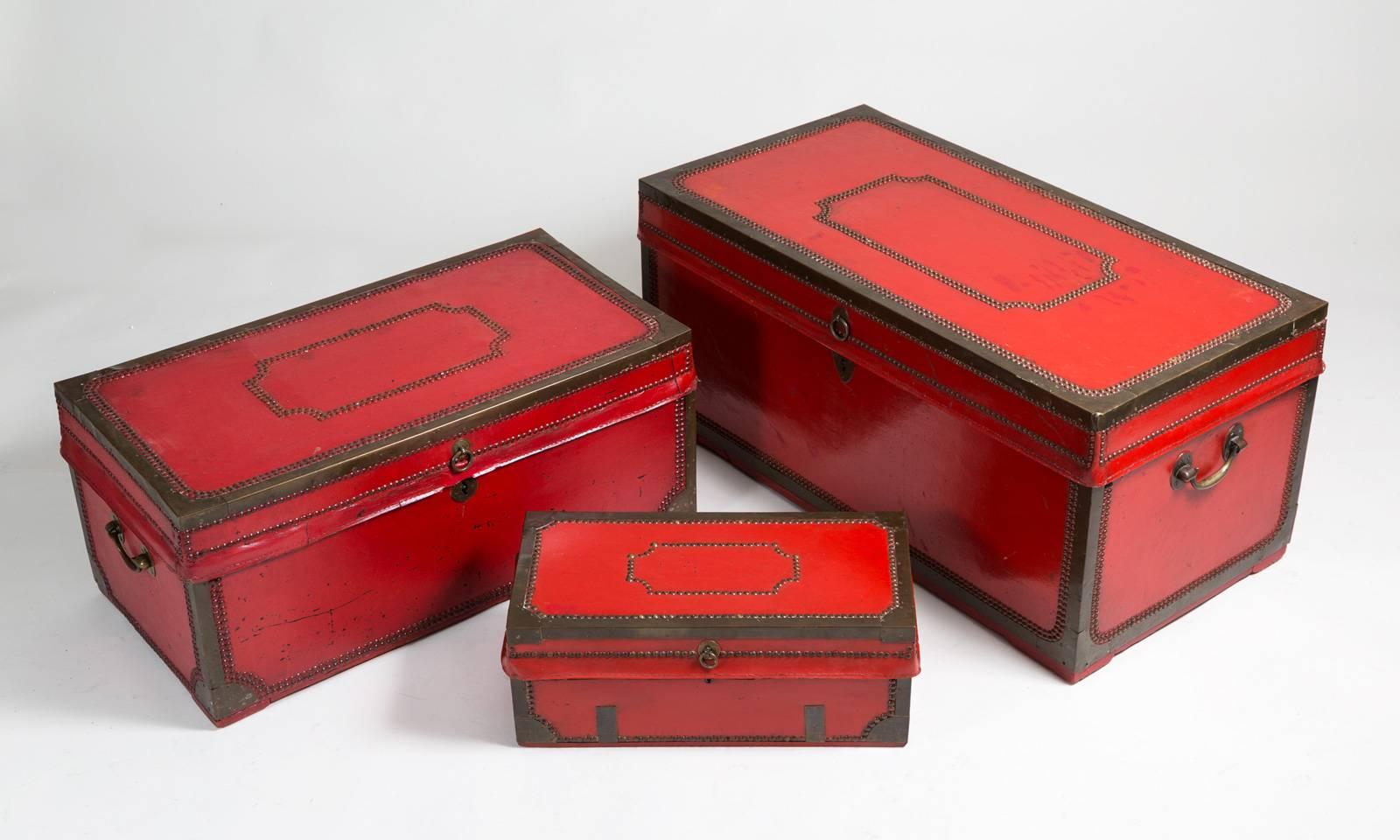 Set of three Chinese export red lacquered leather and brass bound camphor wood trunks, the tops decorated with brass nailheads in shield shapes with brass carrying handles on the sides. They all fit inside each other. The largest trunk’s