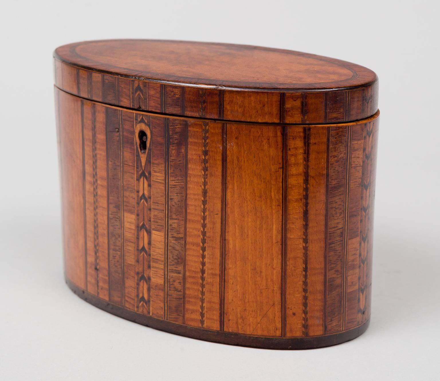 George III oval tea caddy inlaid with specimen woods, the hinged top is mahogany crossbanded with rosewood, the body is decorated with vertical panels of satinwood, rosewood, pear wood and lines of inlay in arrow and checkerboard patterns with ebony