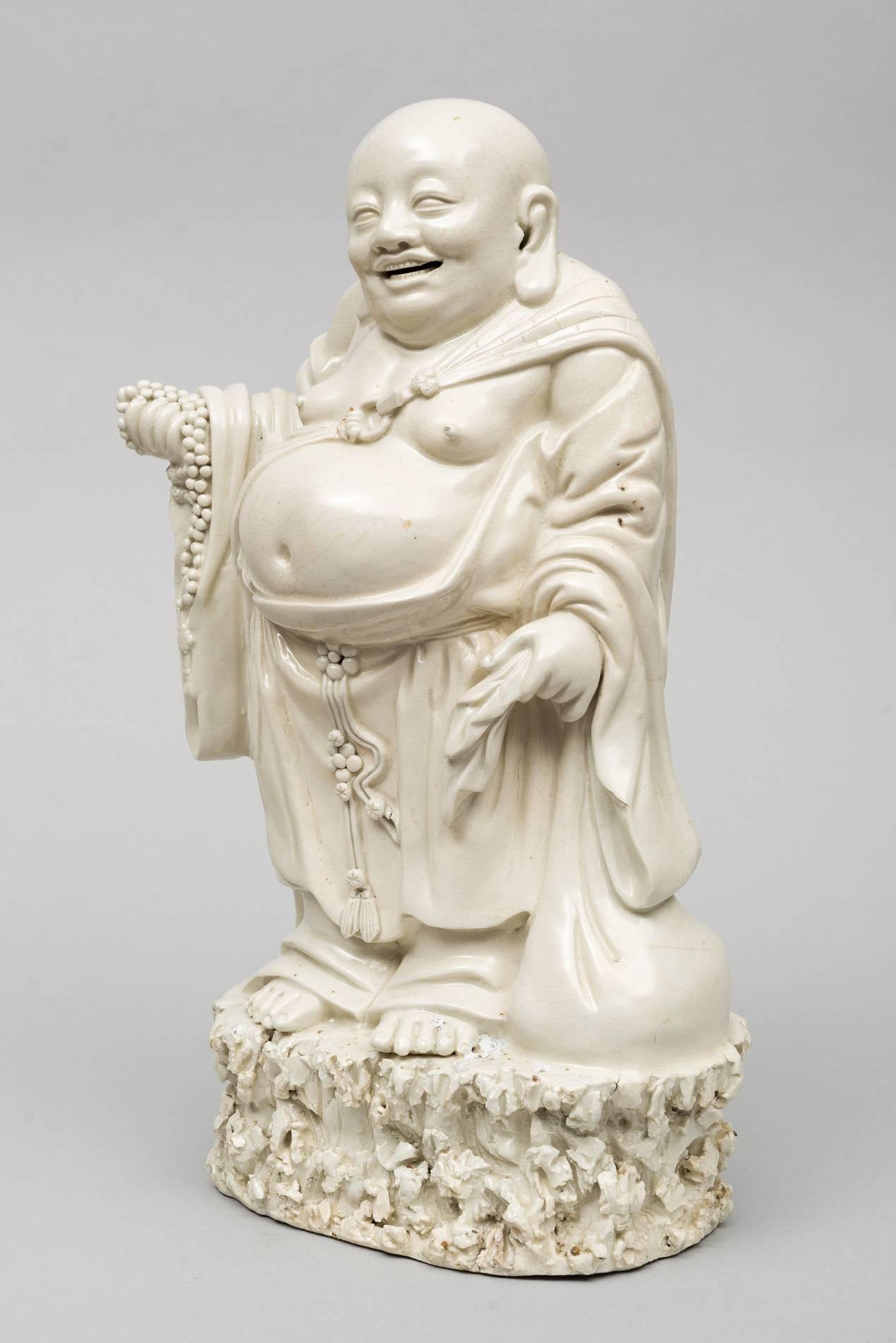 Chinese Blanc de Chine Dehua porcelain Budai or Pu-tai with an open-mouthed smile, holding prayer beads and cloth sack standing on a grassy mount. He is usually identified with or seen as an incarnation of Maitreya, the future Buddha. He is almost