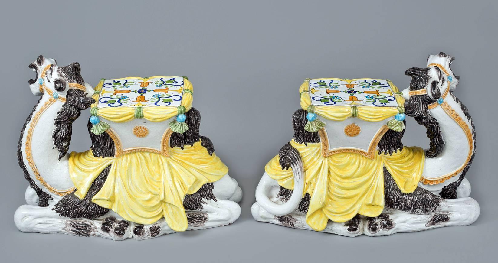 Charming and highly amusing pair of Italian majolica sitting two hump camel side table or garden seat. Between its humps is a saddle decorated with stylized flowers and foliage and draped fabric tied with tassels. Its back is draped with a yellow
